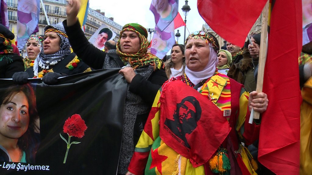 L'image montre une manifestation avec des personnes brandissant des drapeaux et des bannières. Certaines femmes portent des vêtements traditionnels et des accessoires colorés, et elles expriment leurs revendications avec des gestuelles passionnées. Il y a également des portraits visibles en arrière-plan, suggérant un hommage à des individus particuliers. L'ambiance semble être à la fois festive et engagée, avec une forte symbolique culturelle.
