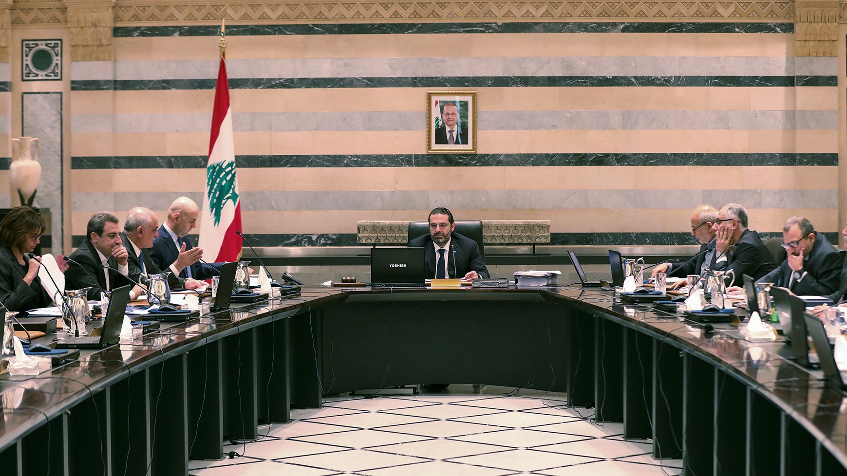 L'image montre une réunion dans une salle de conférence. Au centre, une grande table ovale est entourée de plusieurs personnes vêtues de costumes et cravates. À l'arrière-plan, on peut voir un drapeau libanais et un portrait, probablement du président du Liban, accroché au mur. Les participants semblent être à l'écoute et engagés dans la discussion, avec des documents et des ordinateurs portables devant eux. L'ambiance semble formelle et professionnelle.