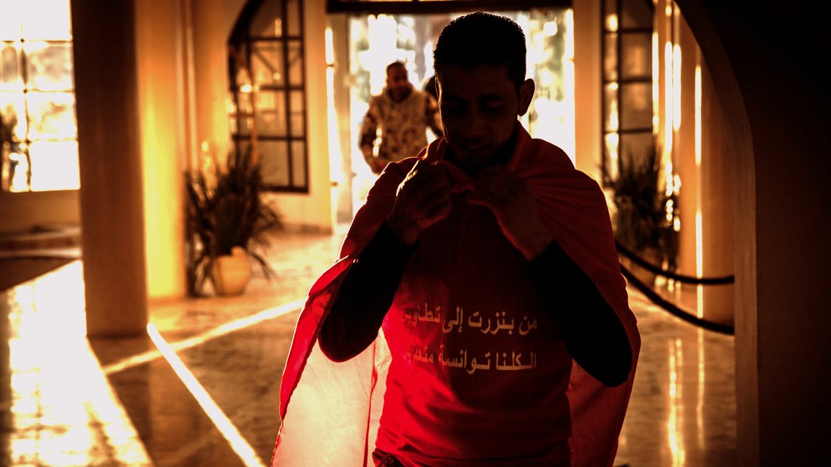 L'image montre un homme portant un t-shirt rouge avec un drapeau. Il semble s'apprêter à sortir, avec une ambiance chaleureuse et lumineuse en arrière-plan. On peut voir des éléments de décor qui suggèrent un espace intérieur, avec des silhouettes floues et une lumière dorée qui crée une atmosphère dramatique. L'homme porte une expression sérieuse et déterminée.