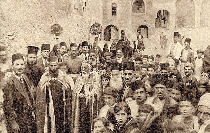 L'image montre un groupe de personnes rassemblées dans une ruelle étroite, probablement d'une ville historique. Au premier plan, on peut voir des hommes en habits traditionnels, certains portant des chapeaux typiques. Un homme central semble être un dignitaire religieux, accompagné d'une procession. En arrière-plan, des enfants et d'autres adultes regardent vers l'objectif, créant une atmosphère de communauté et de partage. Les bâtiments en pierre autour d'eux suggèrent un contexte ancien.