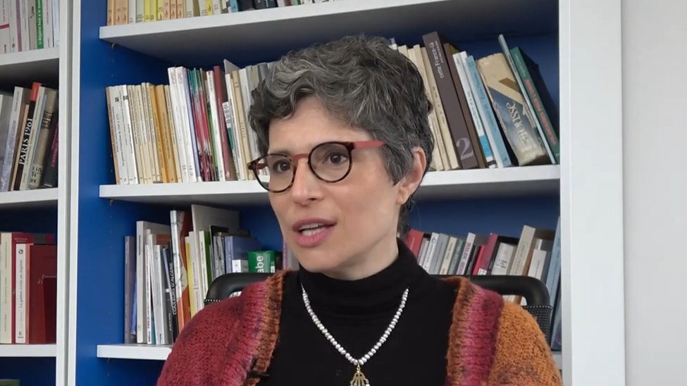 L'image montre une femme avec des cheveux courts et grisonnants, portant des lunettes et un pull noir. Elle est assise devant une bibliothèque remplie de livres, avec un mur bleu derrière elle. Elle a un collier en perles et semble être en train de parler ou de répondre à une question, affichant une expression réfléchie.