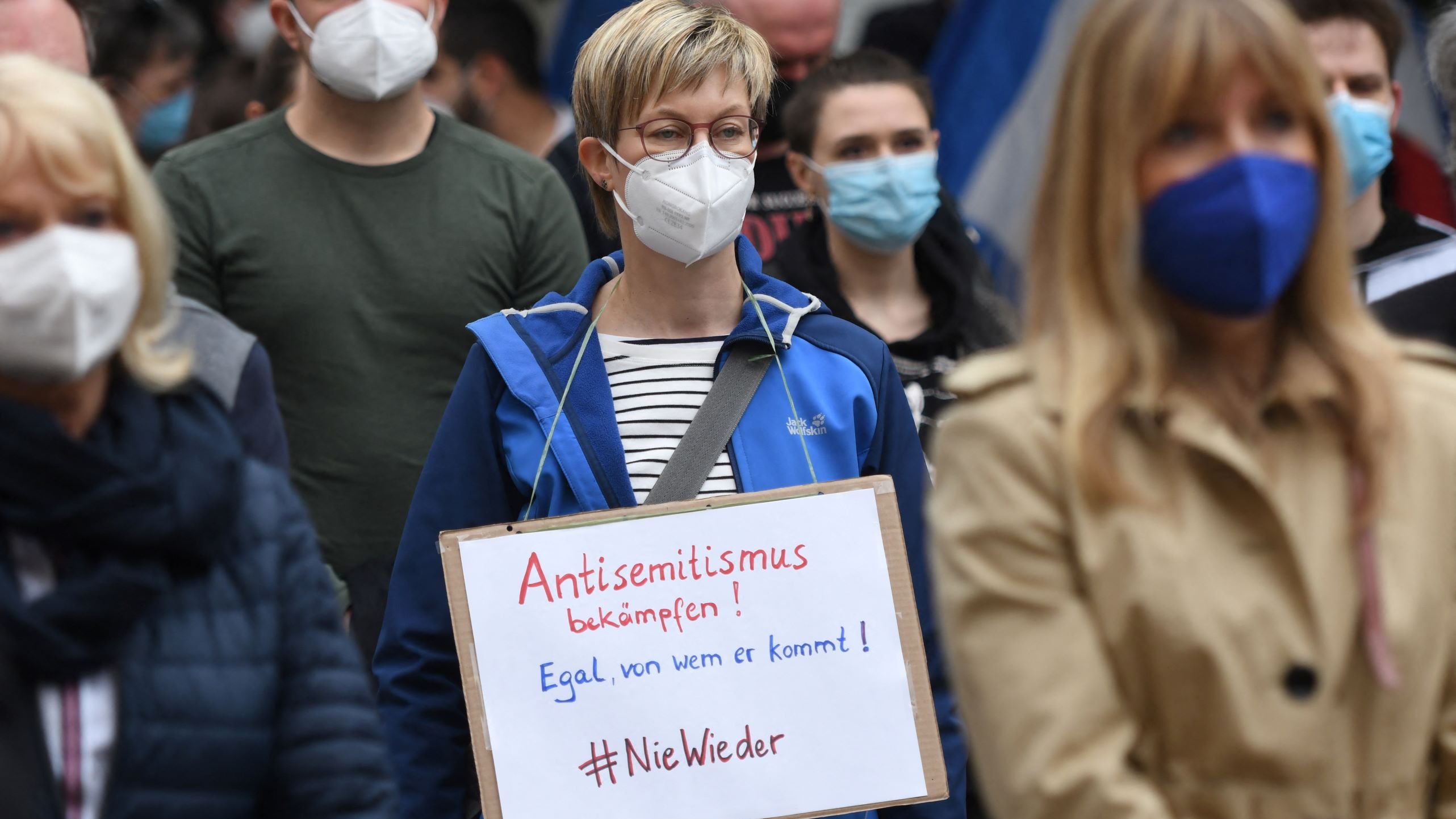 L'image montre une manifestation où des personnes portent des masques. Au premier plan, une femme se distingue avec une pancarte sur laquelle il est écrit en allemand "Antisemitismus bekämpfen! Egal, von wem er kommt! #NieWieder". Elle porte une veste bleue et se tient parmi d'autres manifestants, tous masqués. L'ambiance semble sérieuse et engagée en faveur de la lutte contre l'antisémitisme.