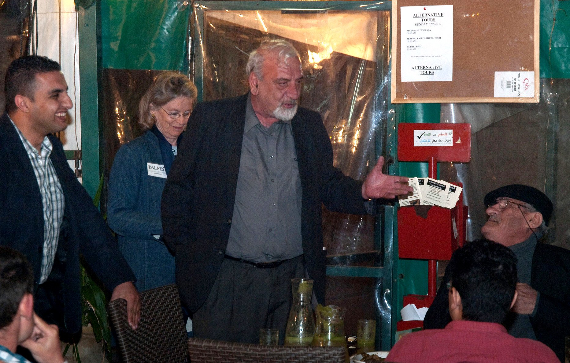 L'image montre un groupe de personnes rassemblées dans un espace qui semble être un restaurant ou une salle de réunion. Un homme au centre, avec des cheveux gris et portant une chemise noire, semble être en train de parler ou de gesticuler. À sa gauche, une femme avec des cheveux ondulés est debout et regardent vers lui. Un autre homme, assis, écoute attentivement, tandis que d'autres personnes sont également présentes à la table. L'environnement est décontracté, avec des éléments typiques de la restauration, tels que des verres et des boissons sur la table.