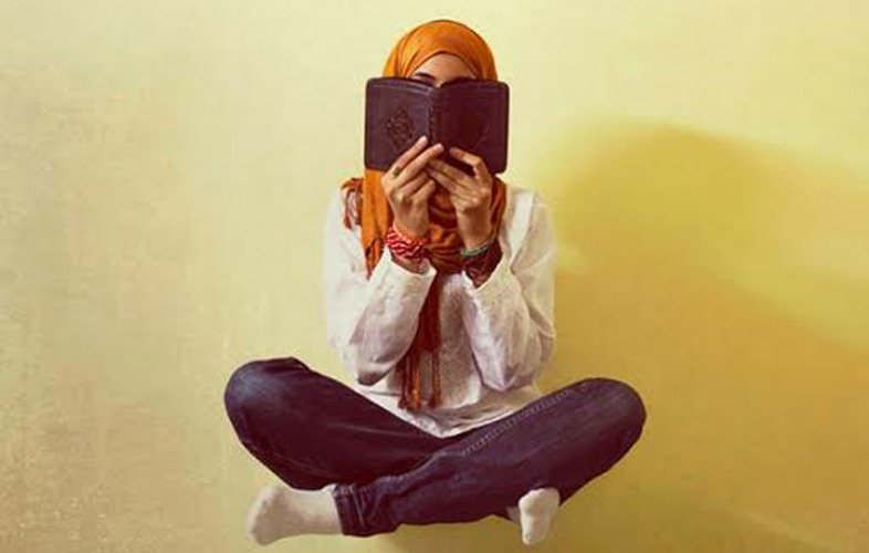 L'image montre une personne assise en tailleur, tenant un livre près de son visage, cachant ainsi son identité. Elle porte un hijab orange et une tenue simple, avec des jeans. L'arrière-plan est d'une couleur claire, créant une ambiance douce et calme. Les bras sont repliés et des bracelets colorés ornent ses poignets. Cette image évoque un moment de réflexion ou de lecture.