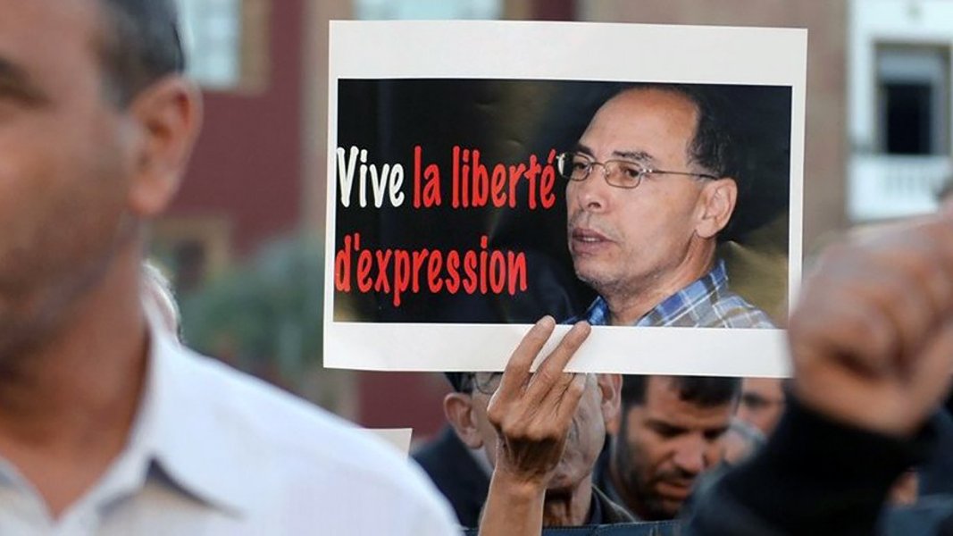 L'image montre une manifestation avec des personnes tenant des pancartes. L'une des pancartes porte le message « Vive la liberté d'expression » en rouge et noir, accompagnée d'une photo d'un homme. L'atmosphère semble engagée et déterminée, reflétant une revendication pour la liberté d'expression. Les manifestants semblent concentrés sur cette cause.