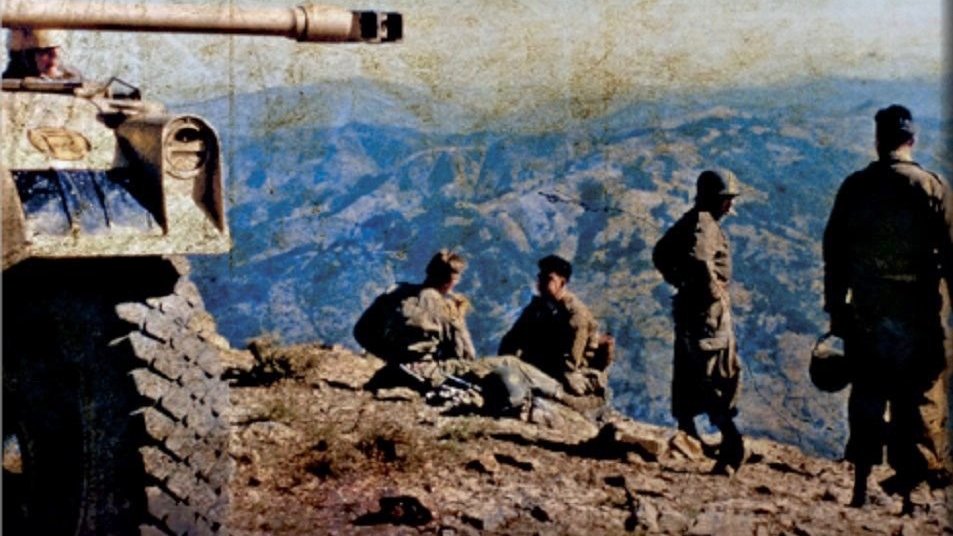 L'image montre un groupe de soldats situés sur un sommet de montagne. À gauche, un char de combat est visible, sa tourelle tournée vers l'avant. Les soldats sont en uniformes militaires, certains semblent assis ou se tiennent debout, avec des paysages montagneux s'étendant en arrière-plan. L'environnement paraît aride, et la lumière donne une atmosphère de calme malgré la présence militaire. Les couleurs de l'image évoquent une certaine nostalgie, suggérant qu'elle pourrait provenir d'une période passée, peut-être de la guerre.