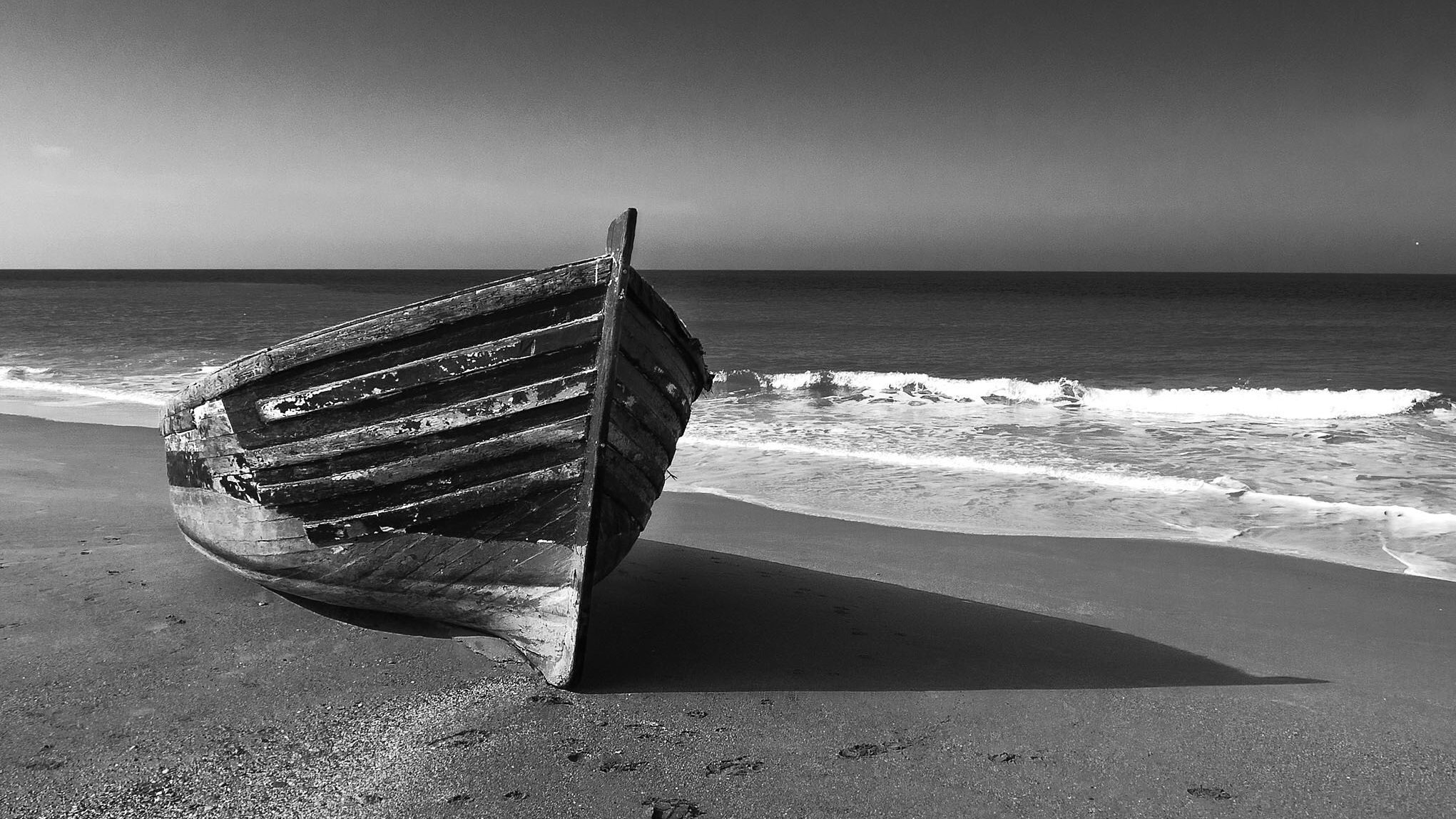 L'image montre un bateau en bois qui est échoué sur une plage. Le bateau, usé par le temps, est orienté vers la mer. La plage est accompagnée de vagues qui se brisent doucement sur le rivage. Le contraste en noir et blanc donne une atmosphère calme et mélancolique à la scène, mettant en valeur les textures du bois et les formes des vagues. Le ciel est dégagé, ajoutant à la sérénité de l'image.