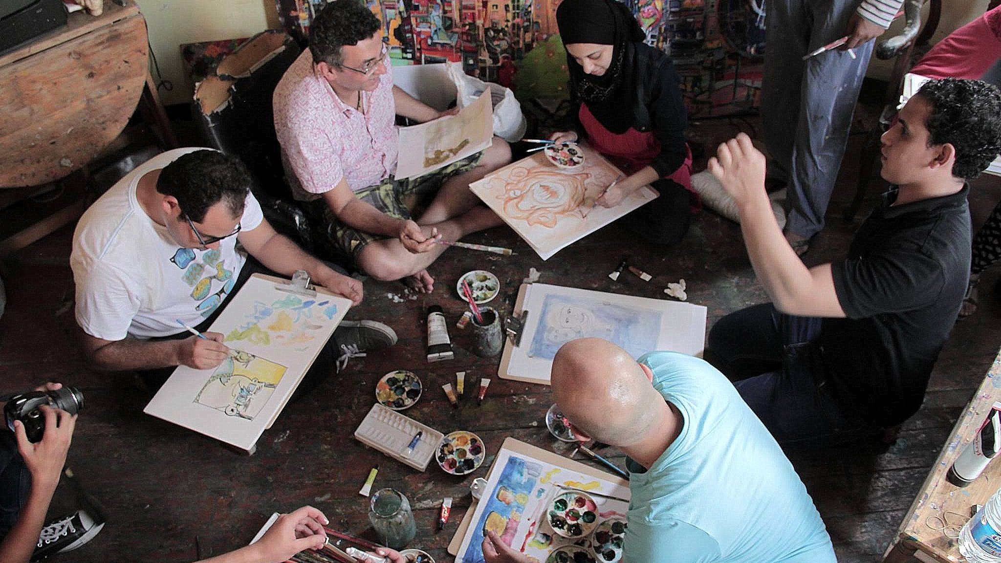 L'image montre un groupe de personnes assises sur le sol dans un espace créatif. Elles semblent engagées dans une activité artistique, utilisant des pinceaux et de la peinture sur des feuilles de papier. Certains participants semblent discuter ou partager des idées, tandis que d'autres se concentrent sur leur travail. L'environnement est coloré, avec des œuvres d'art visibles sur les murs et une palette de peinture au centre. L'ambiance paraît conviviale et collaborative.