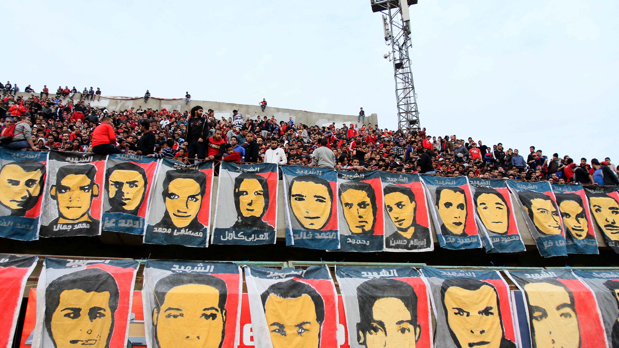 L'image montre un grand groupe de supporters dans un stade, rassemblés dans les gradins. Ils sont entourés de nombreuses banderoles affichant des portraits de différentes personnes, probablement des joueurs ou des figures importantes liés à l'équipe. L'ambiance semble festive et engagée, avec les fans montrant leur passion pour leur équipe. Le ciel est nuageux et le stade est bien rempli de spectateurs.
