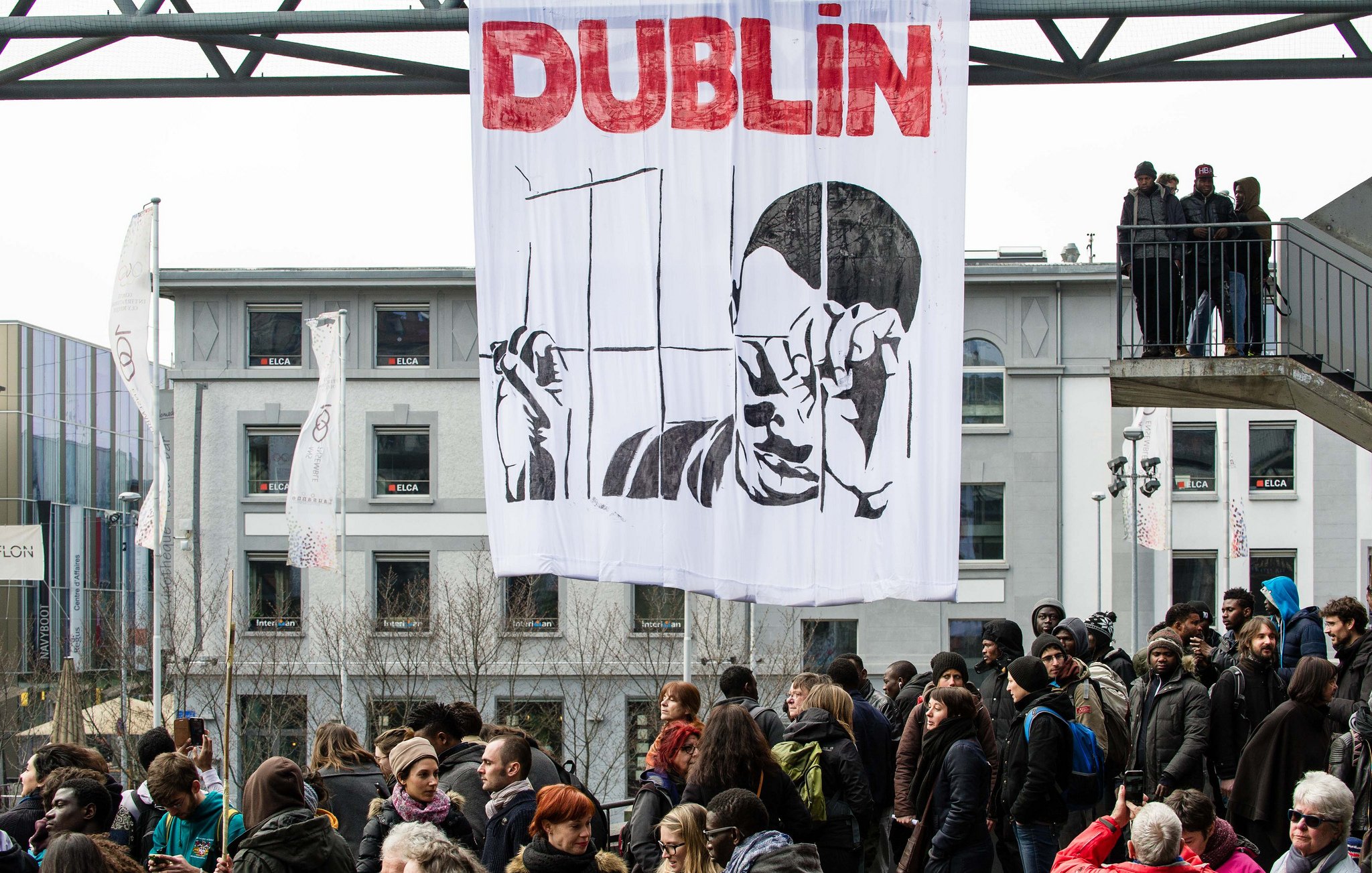 L'image montre une manifestation à Dublin, avec une grande banderole suspendue qui représente une figure tenant des barres comme une prison. En arrière-plan, on peut voir un groupe de personnes rassemblées, manifestant sur une route ou une place. Les gens semblent engagés dans la cause, et l'atmosphère est dynamique, probablement en réponse à une problématique sociale ou politique.
