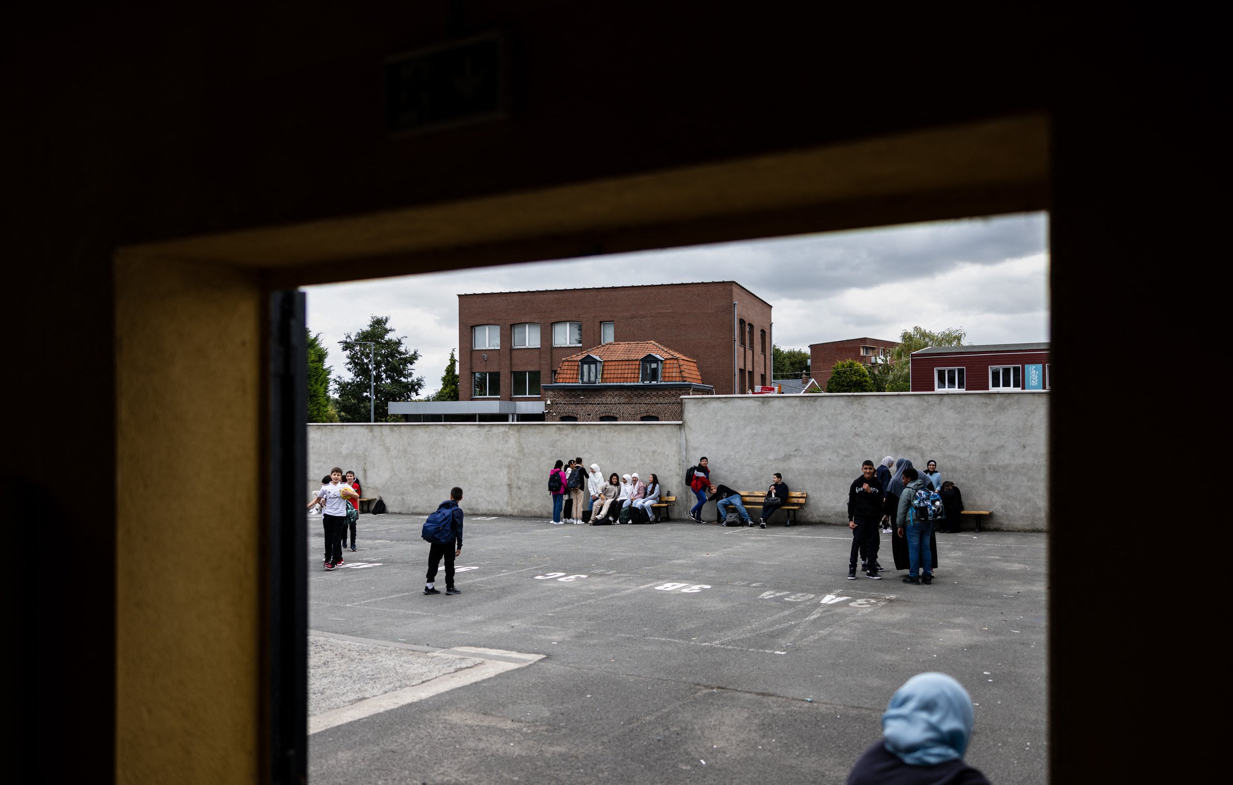L'image montre une scène d'un espace extérieur, probablement une cour de récréation ou un terrain de sport. On peut voir plusieurs groupes de jeunes, répartis sur le sol, assis ou discutant ensemble. À l'arrière-plan, il y a des bâtiments avec des fenêtres et un toit de briques. Le ciel est nuageux, ce qui donne une ambiance un peu sombre à la scène. Les jeunes semblent engagés dans des conversations ou des activités sociales.