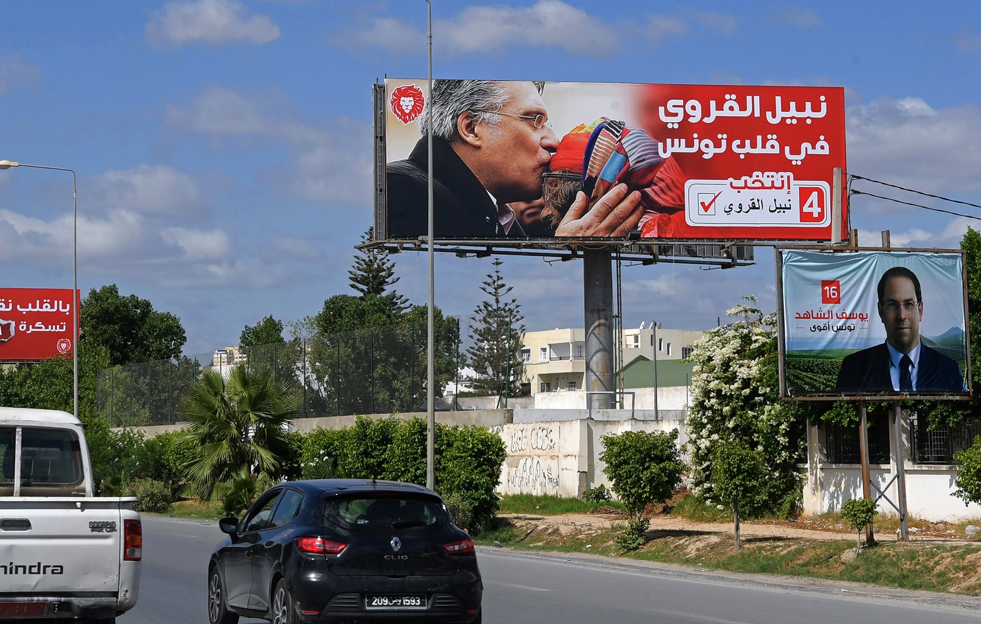 Sur cette image, on voit un panneau publicitaire en arabe. Le panneau montre deux personnages qui s'embrassent, entourés de couleurs vives et de slogans. En bas, on peut voir une photo d'une autre personne (peut-être un candidat) avec un texte supplémentaire à son propos. La scène se déroule dans un environnement urbain, avec des voitures circulant sur une route. Le ciel est bleu, parsemé de nuages.