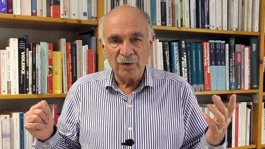 L'image montre un homme debout devant une étagère remplie de livres. Il porte une chemise à rayures et semble s'adresser à un public ou donner une présentation. Ses mains sont levées, ce qui indique qu'il est en train de faire un point ou d'expliquer quelque chose. L'arrière-plan est constitué de livres aux couvertures variées, suggérant un environnement intellectuel ou académique.