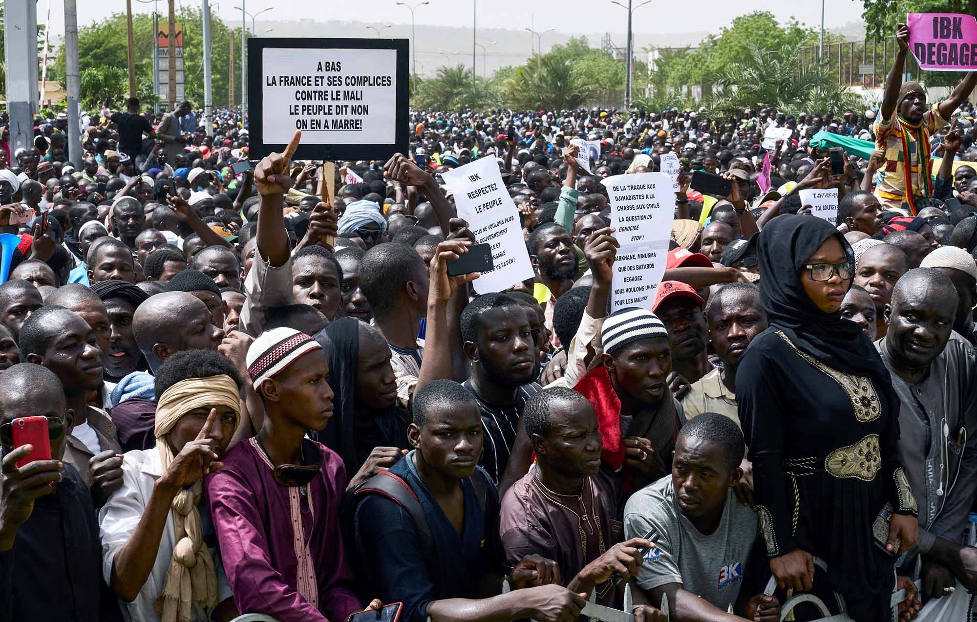 L'image montre une grande foule de manifestants réunis, tenant des pancartes. Les personnes semblent exprimer leur mécontentement et leurs revendications. On peut voir une diversité de vêtements parmi les participants, allant des habits traditionnels à des vêtements plus modernes. L'atmosphère paraît énergique et engagée, avec des individus qui tentent de faire entendre leur voix. Des barrières sont présentes pour délimiter l'espace.