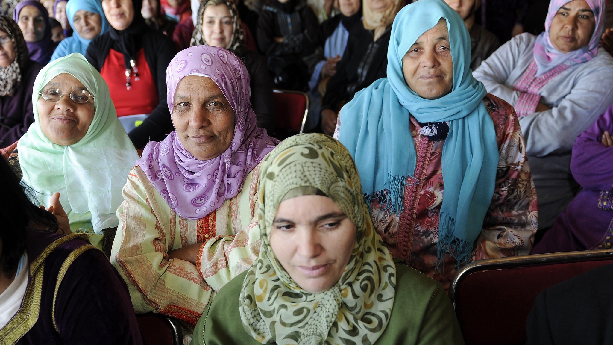 L'image montre un groupe de femmes assises, majoritairement vêtues de vêtements traditionnels et portant des foulards. Certaines portent des vêtements clairs tandis que d'autres ont des tenues plus sombres. Les expressions des femmes varient, allant de souriantes à attentives. L'environnement semble être une réunion ou un rassemblement, avec des murs ornés de motifs. L'atmosphère semble conviviale et solennelle.