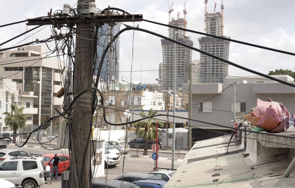 L'image montre une vue urbaine où plusieurs poteaux électriques sont entremêlés de fils. À l'arrière-plan, on peut apercevoir deux grands immeubles en construction. Au premier plan, il y a des bâtiments plus petits et des véhicules stationnés. On peut également voir des palmiers, ce qui indique un environnement peut-être chaud ou méditerranéen. L'ensemble donne une impression de ville en développement, avec un mélange de structures anciennes et modernes.