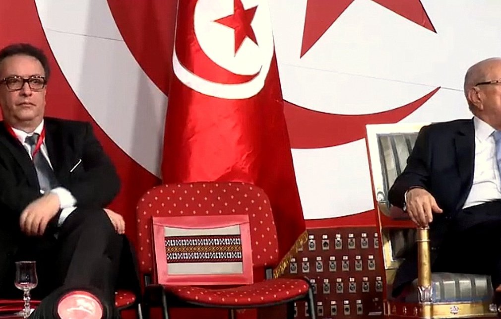 L'image montre deux hommes assis sur des chaises lors d'un événement. Ils semblent être en train d'écouter ou d'observer quelque chose, peut-être un discours ou une présentation. En arrière-plan, il y a un drapeau tunisien, ce qui indique que l'événement se déroule en Tunisie. L'un des hommes porte des lunettes et a une expression sérieuse, tandis que l'autre exerce une posture détendue. La scène semble officielle, peut-être liée à la politique ou à un rassemblement important.