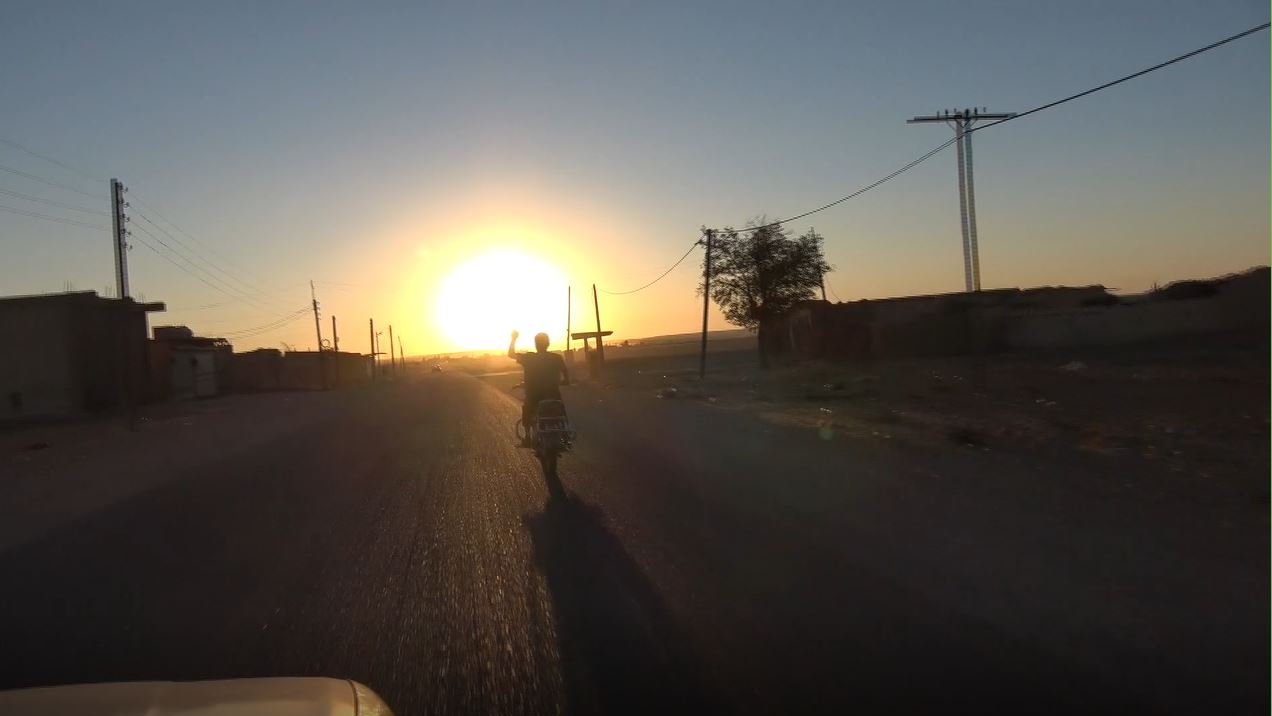 L'image montre un paysage rural au crépuscule. On voit une route désertique s'étendant devant, avec un motard roulant vers la direction du soleil couchant. Les rayons du soleil illuminent le ciel, créant une ambiance chaleureuse. De chaque côté de la route, on aperçoit quelques bâtiments et des poteaux électriques. L'atmosphère semble paisible et tranquille.