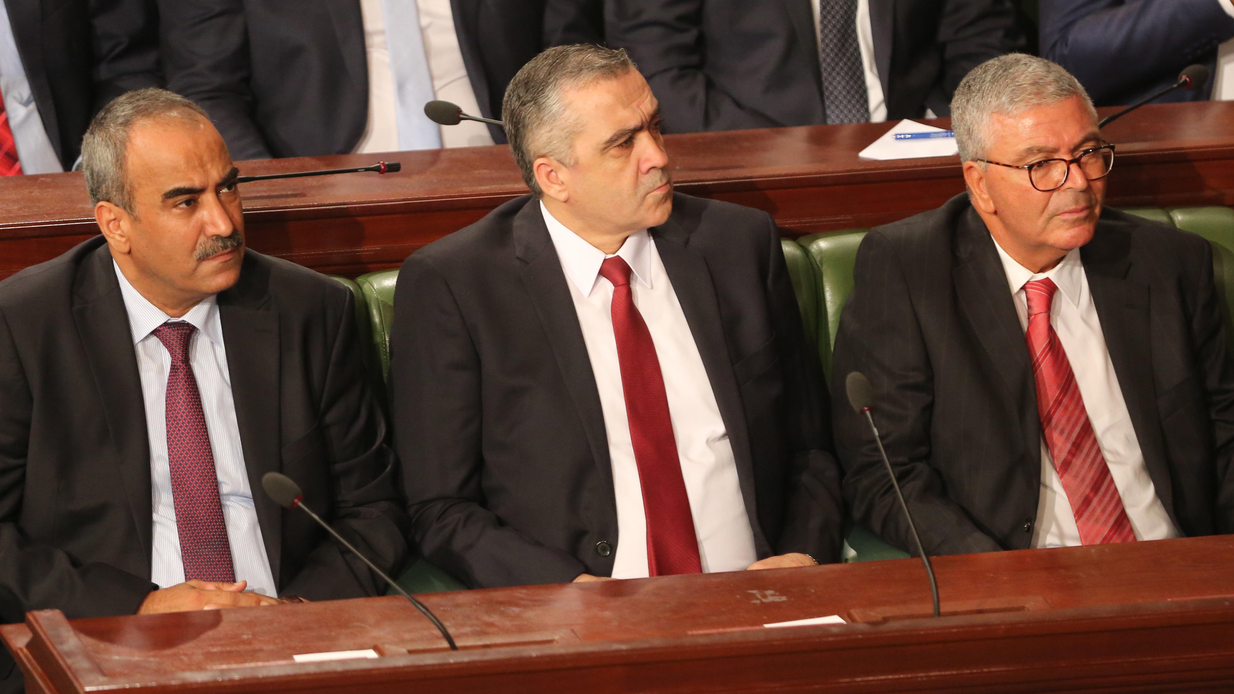 L'image montre trois hommes assis côte à côte dans un cadre formel, probablement lors d'une réunion ou d'une séance parlementaire. Ils portent des costumes sombres et des cravates, affichant une expression sérieuse. L'un d'eux porte une cravate rouge, tandis que les autres optent pour des nuances plus sombres. En arrière-plan, on aperçoit des personnes en costume, suggérant un environnement officiel.