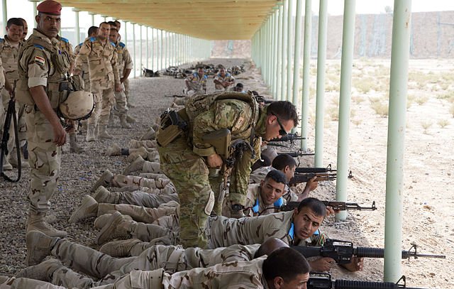 L'image montre un groupe de soldats en train de s'entraîner sur un champ de tir, allongés à terre avec leurs armes. Certains soldats portent des uniformes beiges avec des motifs camouflés, tandis qu'un instructeur semble donner des conseils à un autre soldat. En arrière-plan, on aperçoit d'autres soldats attendant ou surveillant l'exercice. L'environnement est désertique avec une structure en métal au-dessus d'eux, offrant un abri contre le soleil.