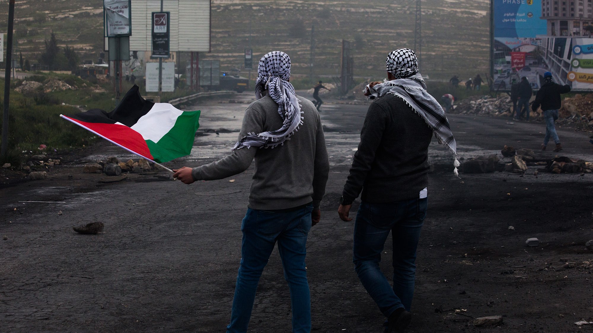 L'image montre deux hommes marchant sur une route, chacun portant un keffieh. L'un d'eux tient un drapeau palestinien, qui est composé de bandes noire, blanche, verte et rouge. L'arrière-plan présente des panneaux publicitaires, des débris et une ambiance brumeuse. Les hommes semblent se diriger vers une direction, symbolisant probablement une manifestation ou une action de revendication.