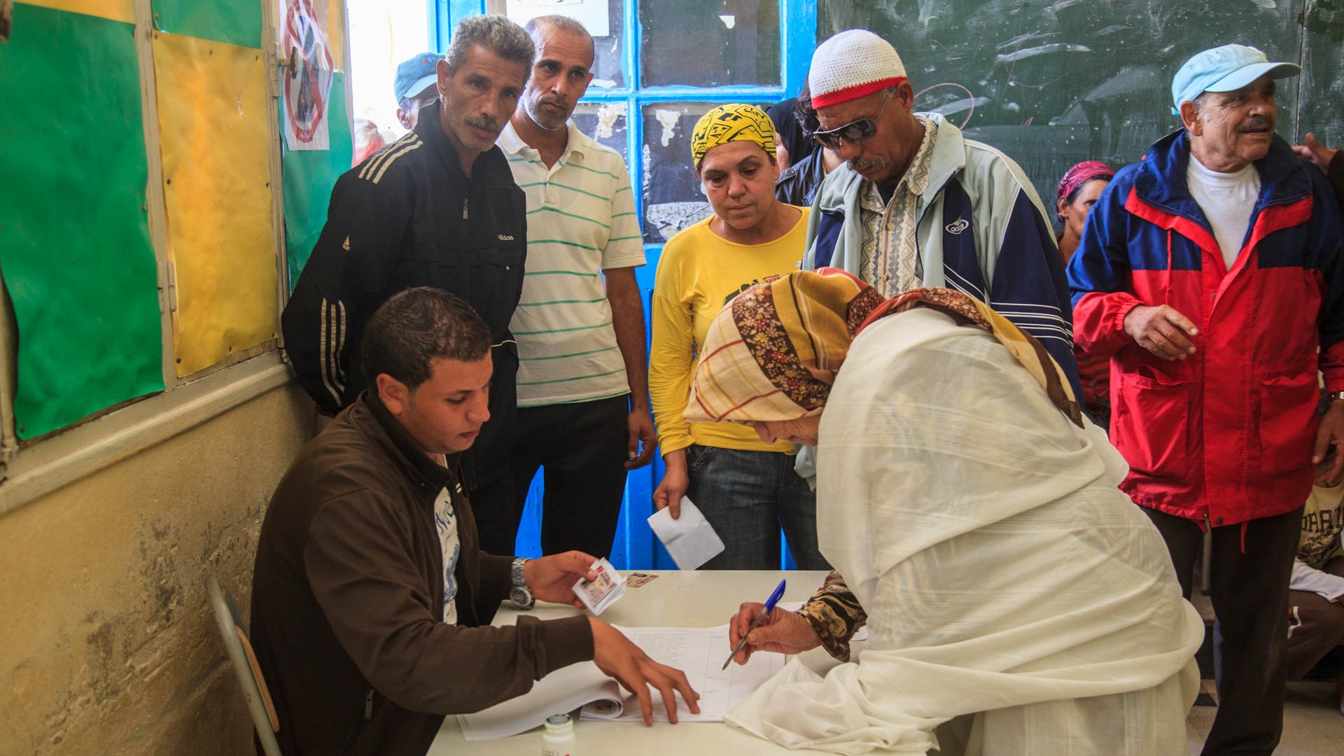 L'image montre un groupe de personnes se rassemblant autour d'un bureau dans une salle. Un homme est assis à une table, où il semble aider une femme, qui est en train de signer un document. Elle porte une tenue claire et un foulard. D'autres personnes, dont des hommes et des femmes, observent la scène, manifestant un intérêt. Les murs sont décorés de couleurs vives, et une fenêtre ouverte laisse entrer la lumière. L'environnement semble informer d'une activité communautaire, peut-être liée à un vote ou à une inscription.