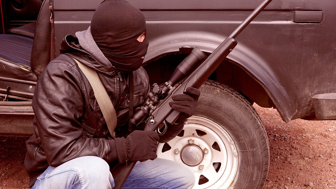 L'image représente une personne en position accroupie, portant un masque noir qui couvre son visage. Elle est habillée d'une veste en cuir et de gants noirs. Dans ses mains, elle tient un fusil équipé d'un accessoire de visée. En arrière-plan, on peut voir une camionnette avec une roue visible, suggérant un environnement extérieur. L'ensemble dégage une atmosphère tendue et dramatique.