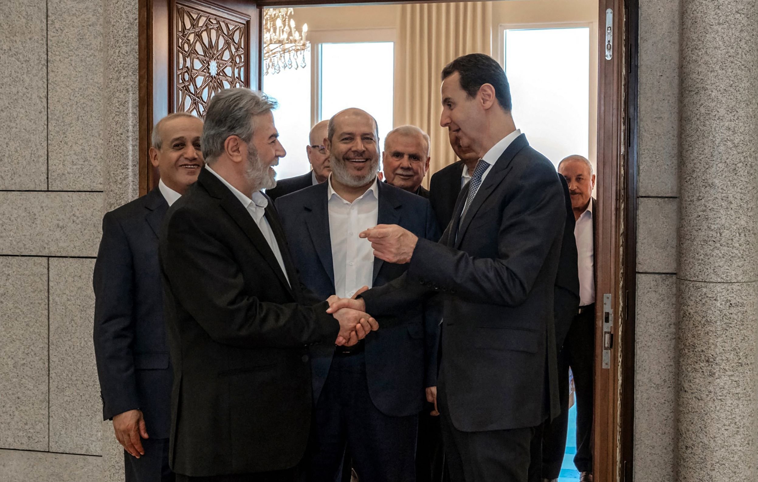 L'image montre un groupe d'hommes en train de se rencontrer dans un intérieur. Plusieurs d'entre eux sourient et semblent engagés dans une conversation amicale. L'un des hommes, au premier plan, serre la main d'un autre, tandis que les autres les observent. L'environnement semble formel, avec des éléments décoratifs visibles, comme une porte ornée et des fenêtres laissant entrer la lumière.