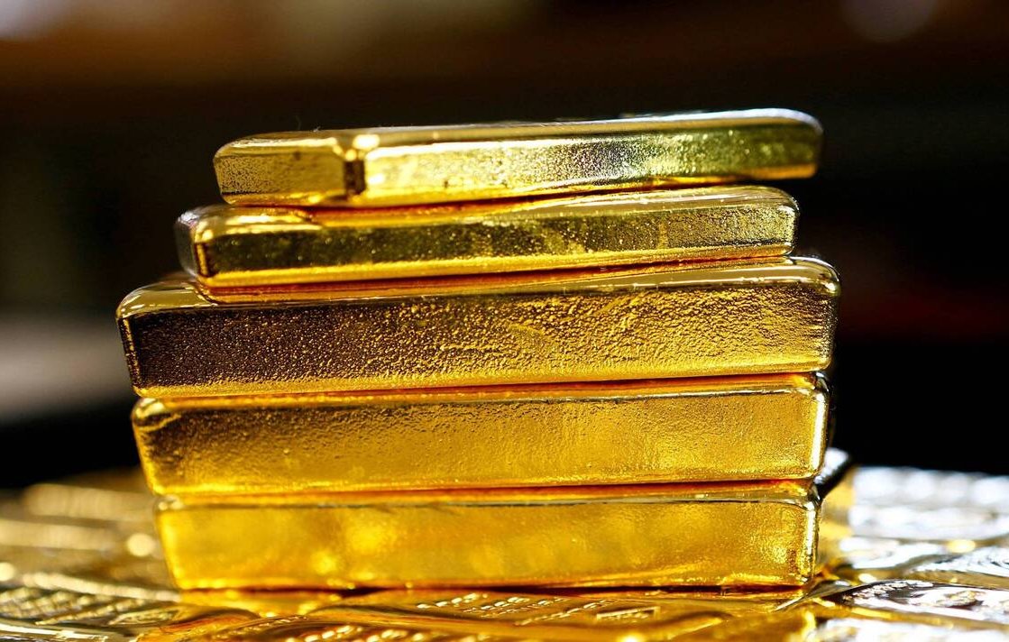 L'image montre plusieurs barres en or empilées les unes sur les autres. Chaque barre a une surface brillante et métallique, affichant la couleur dorée caractéristique de l'or. Les barres semblent bien travaillées, avec des contours nets et une texture réfléchissante. En arrière-plan, on devine une surface moins définie, ce qui met en valeur le brillant des barres d'or au premier plan.