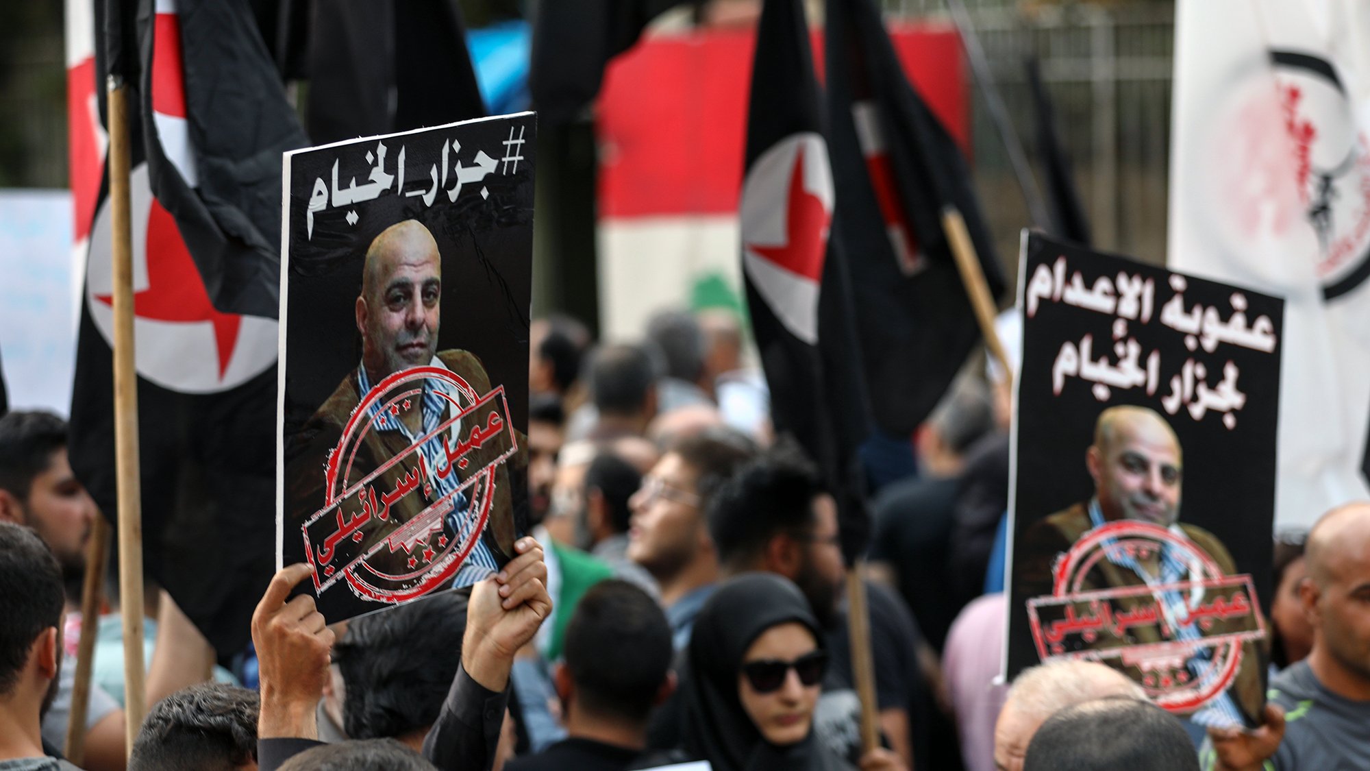 L'image montre une manifestation avec des personnes tenant des pancartes et des drapeaux. Les pancartes contiennent des portraits de personnes et des textes en arabe, évoquant des messages politiques ou sociaux. La foule semble engagée dans une cause, exprimant des émotions fortes. On peut voir une diversité de participants, certains portant des vêtements traditionnels et d'autres plus modernes, ainsi qu'un fond urbain suggérant un rassemblement public.