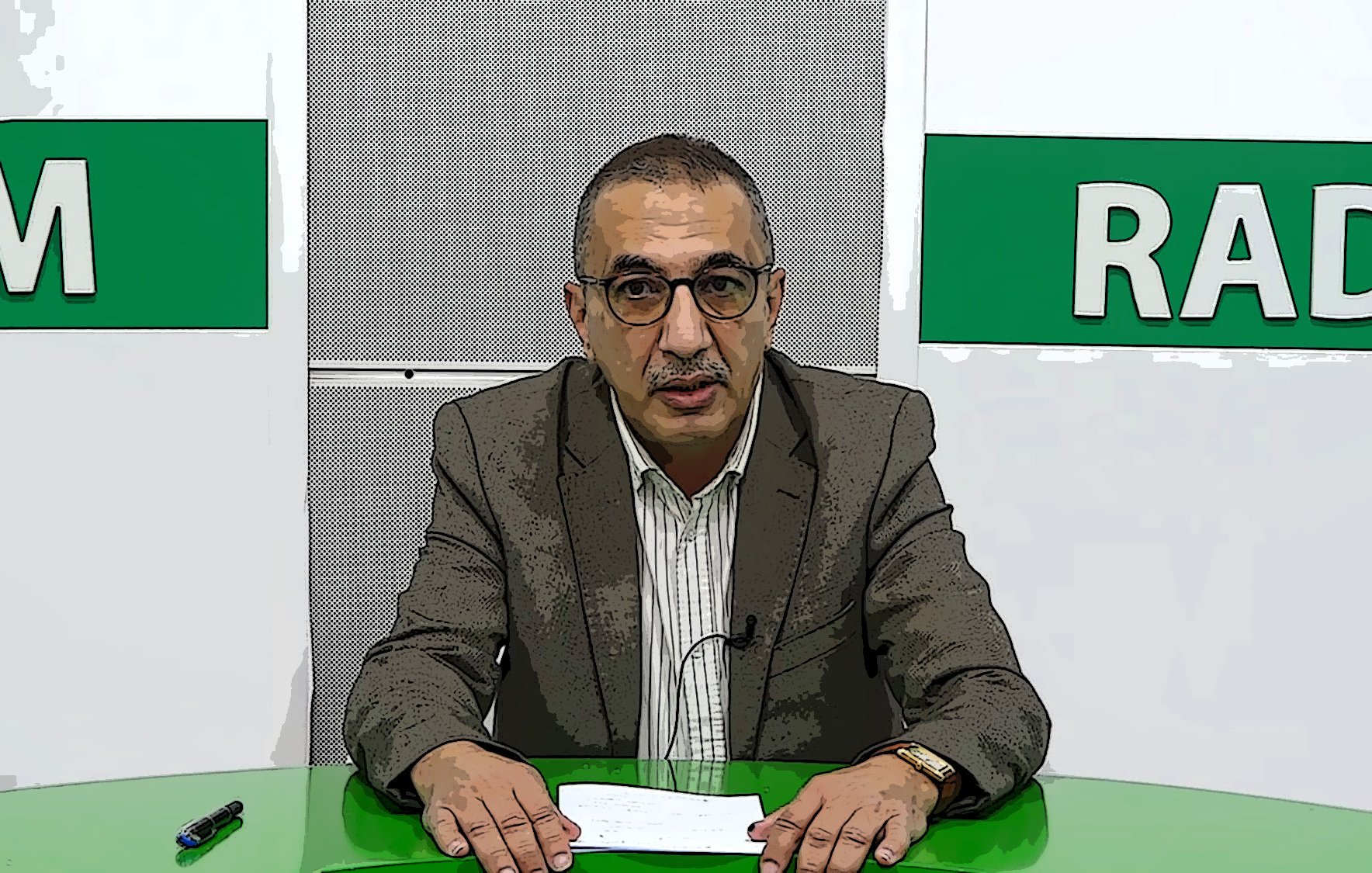 L'image montre un homme devant un bureau, probablement dans un studio de télévision ou de radio. Il porte des lunettes et un costume, avec une chemise rayée. Le bureau est vert, et il semble tenir des documents ou un script. En arrière-plan, il y a des éléments graphiques avec les mots "RADIO" et une autre inscription non visible. L'homme a une expression sérieuse et semble prêt à faire une présentation ou une annonce.