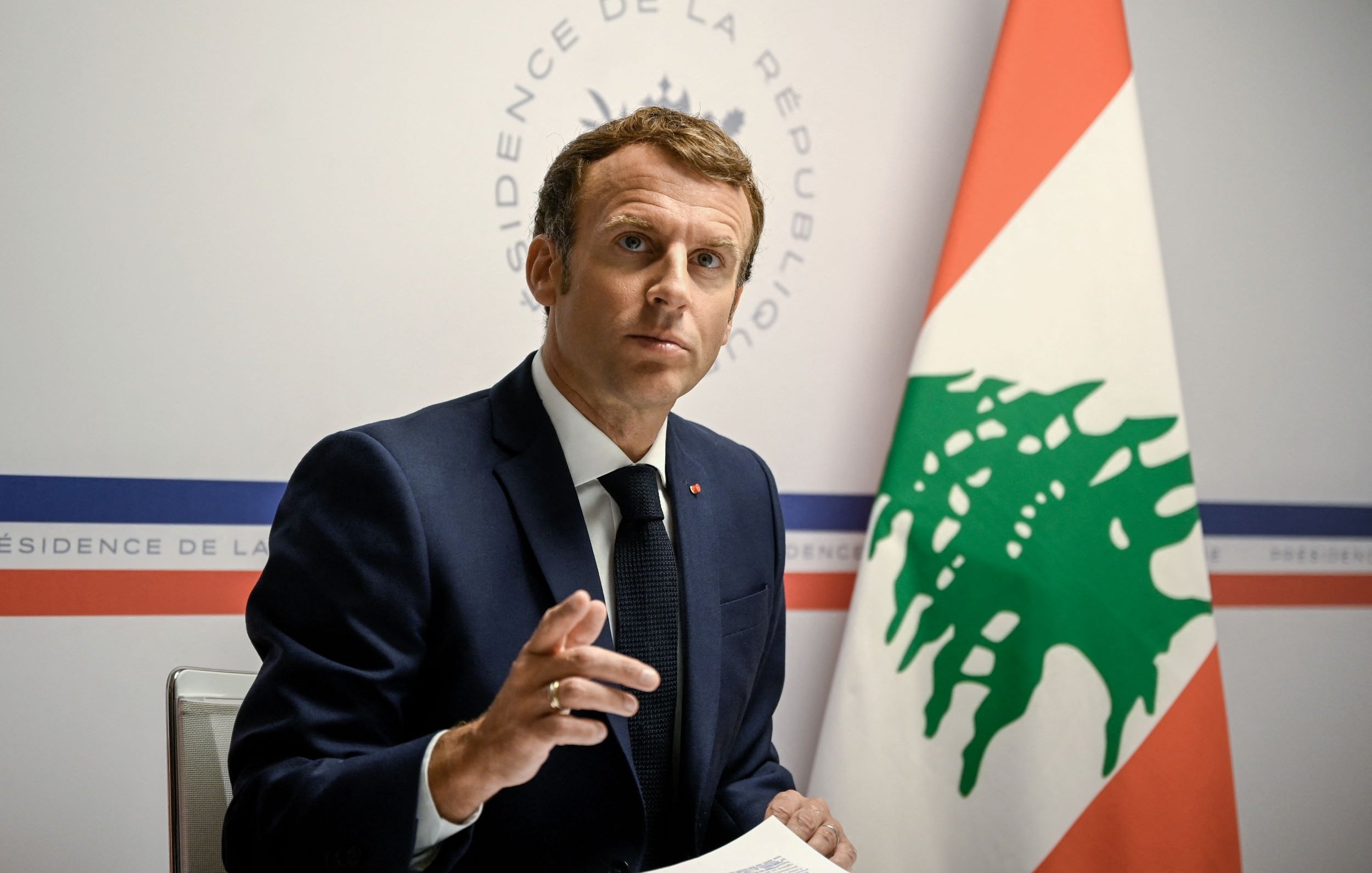 L'image montre un homme en costume, assis à une table, s'adressant à un public. Il semble être en train de faire un discours ou de répondre à des questions, avec une expression sérieuse. En arrière-plan, on peut voir un drapeau libanais ainsi qu'un logo indiquant "Présidence de la République". L'ambiance de l'image évoque un contexte officiel et diplomatique.