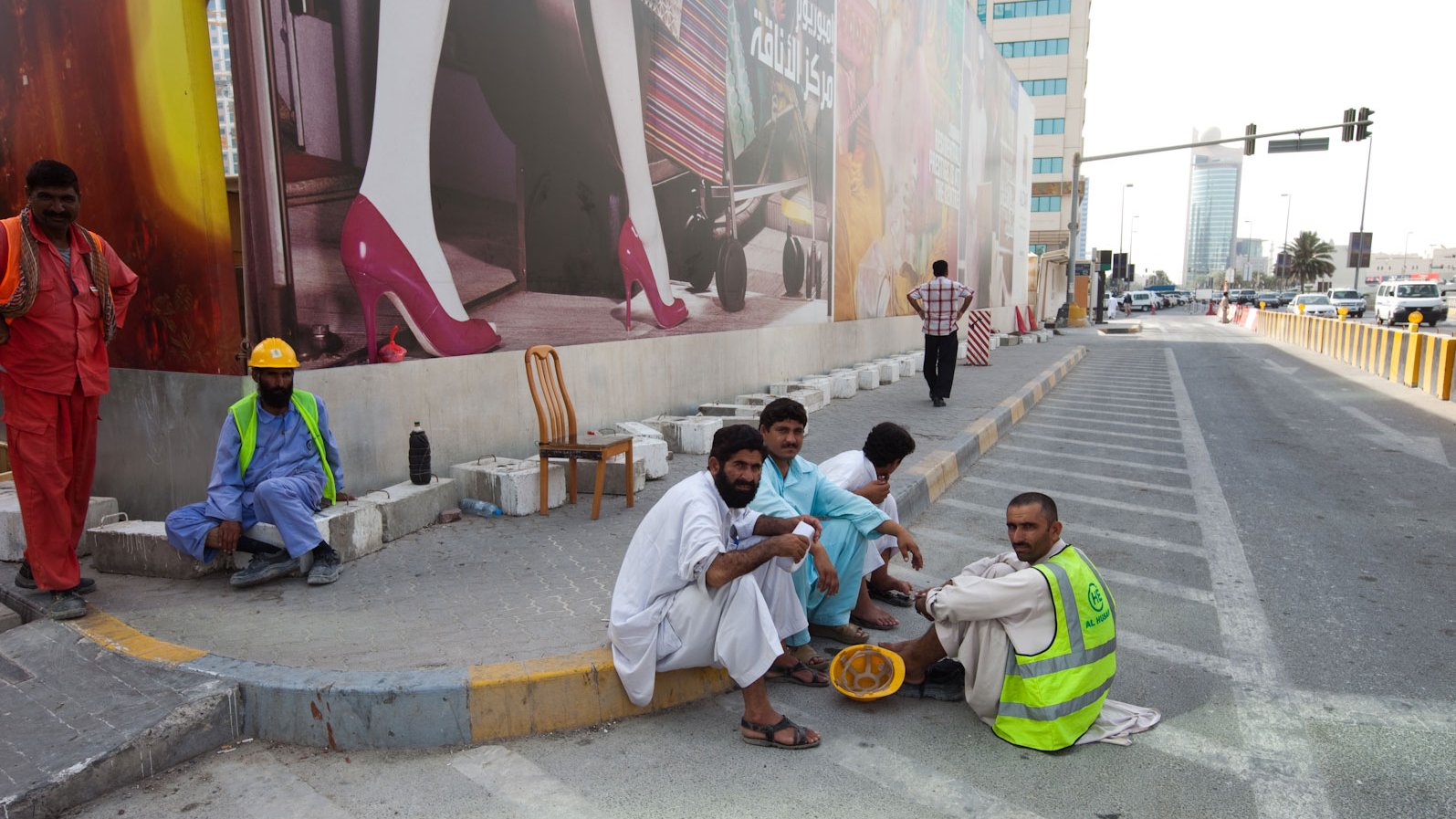 L'image montre un groupe d'hommes assis sur un trottoir, probablement en train de faire une pause. Ils portent des vêtements traditionnels, et certains portent des gilets de travail. En arrière-plan, on aperçoit un grand panneau publicitaire avec des images colorées. L'environnement urbain est visible, avec des bâtiments modernes et une route. L'atmosphère semble calme, et les hommes semblent discuter entre eux.