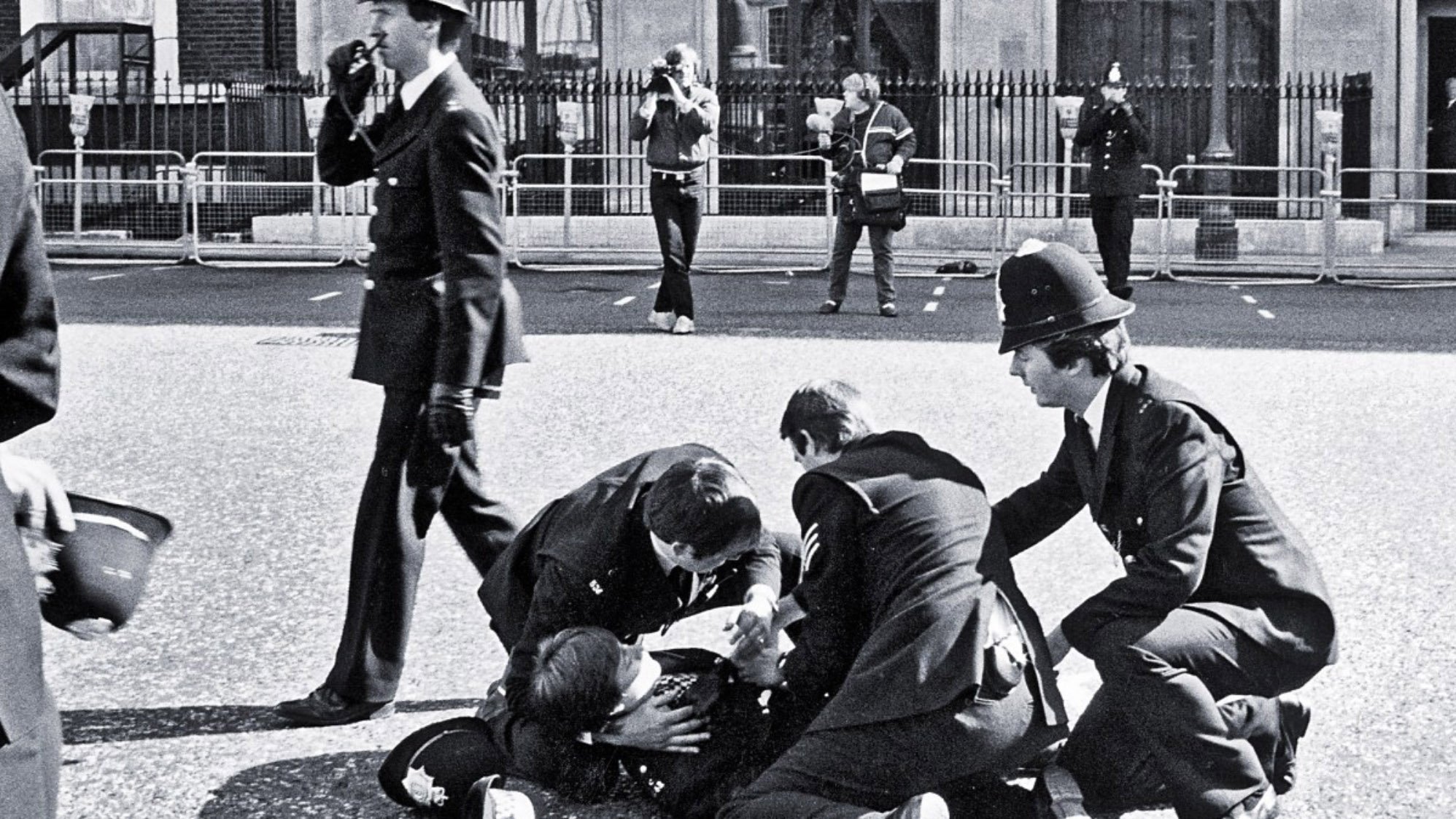 L'image montre une scène policière en noir et blanc. Plusieurs policiers en uniforme entourent une personne au sol, visiblement en train d'être arrêté ou assisté. D'autres policiers se tiennent en arrière-plan, certains prenant des notes ou observant la situation. L'environnement semble urbain, avec des barrières de sécurité visibles, probablement autour d'une zone publique. L'atmosphère dégage une impression d'urgence et d'autorité.