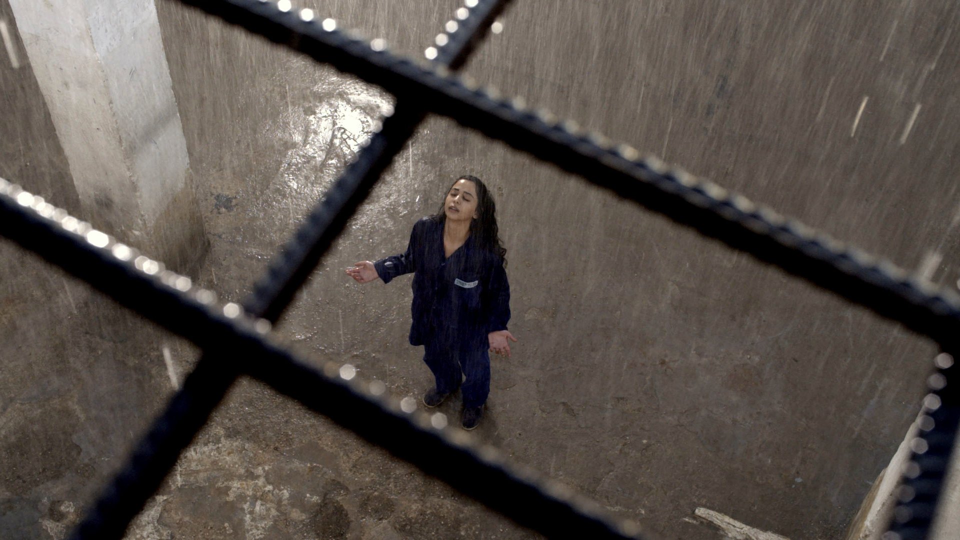 L'image montre une scène dramatique d'une femme debout dans un espace clos, probablement une cellule. Elle porte une tenue de prison et semble être sous une forte pluie qui tombe du plafond. La lumière est tamisée, créant une ambiance sombre et mélancolique. La femme, les bras ouverts et la tête inclinée vers le ciel, exprime une pose de désespoir ou de lâcher-prise, tandis que des barreaux en fer témoignent de son confinement. Le sol semble humide et dégradé, ce qui renforce l'atmosphère oppressante de l'environnement.