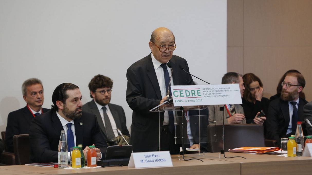 L'image montre un homme âgé, probablement un orateur, se tenant derrière un podium avec un panneau portant le mot "CEDRE" et la date de l'événement. Il semble prendre la parole lors d'une conférence. Autour de lui, plusieurs autres personnes sont assises, attentives, et il y a des bouteilles d'eau et des boissons sur la table. L'ambiance semble formelle et professionnelle, suggérant un événement important.