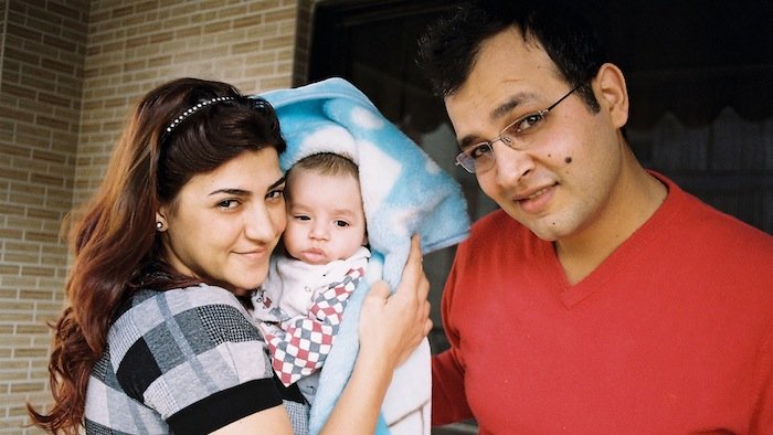 Sur cette image, on voit un couple et un bébé. La femme, à gauche, sourit et tient le bébé enveloppé dans une couverture, tandis qu'un homme, à droite, regarde la caméra en souriant également. Ils se tiennent tous les trois dans un environnement intérieur, probablement chez eux, avec un mur en briques en arrière-plan. L'atmosphère générale est chaleureuse et affectueuse.