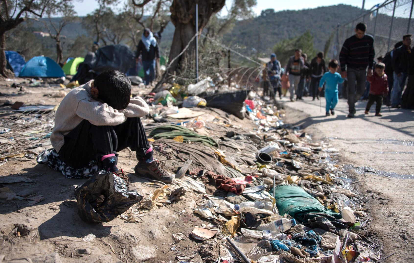 L'image montre un jeune enfant assis au sol, semblant triste ou désemparé. Il est entouré de déchets et de débris, ce qui souligne une situation de pauvreté et de désespoir. À l'arrière-plan, on peut voir d'autres personnes marchant, probablement des familles, dans un environnement difficile. Des tentes sont visibles, indiquant un campement temporaire. L'ensemble de la scène évoque une réalité difficile et poignante.