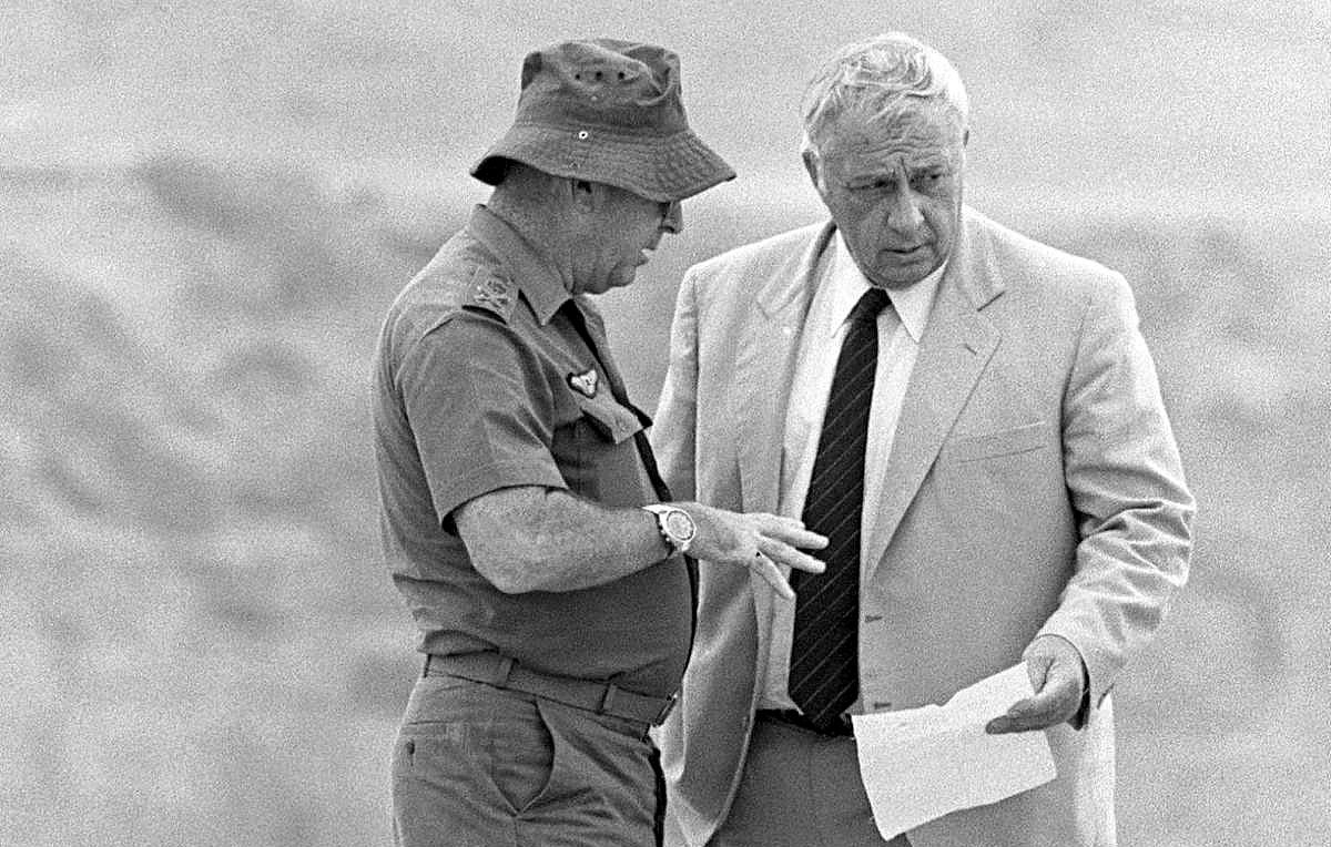 L'image montre deux hommes en train de discuter. L'un d'eux porte une tenue militaire, incluant un chapeau, tandis que l'autre est vêtu d'un costume clair. Ils semblent examiner un document ensemble et échanger des informations de manière sérieuse. L'arrière-plan est flou, suggérant un environnement extérieur. La scène dégage une impression de concentration et d'importance.