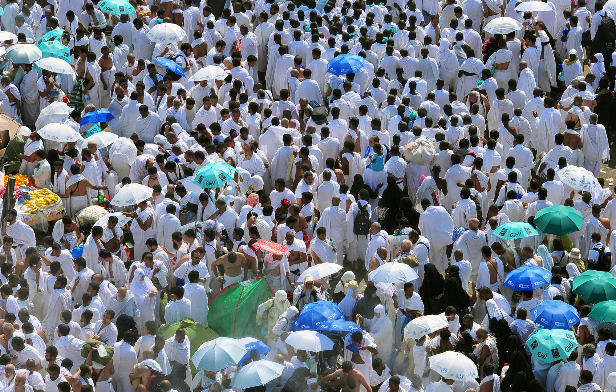 L'image montre une grande foule de personnes vêtues de vêtements blancs, probablement en train de participer à un pèlerinage ou à un événement religieux. De nombreux participants arborent des parapluies colorés, qui offrent de l'ombre dans un environnement ensoleillé. L'atmosphère semble très animée, avec un grand nombre de personnes rassemblées, ce qui reflète un moment de rassemblement spirituel.