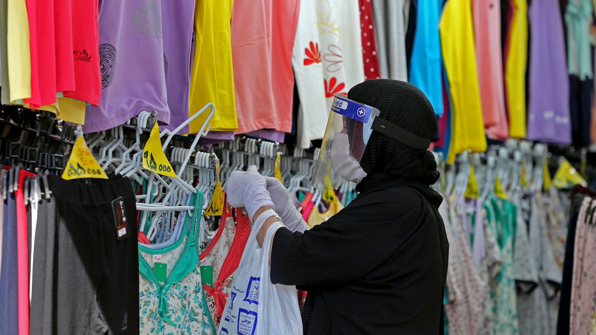 L'image montre une personne dans un magasin de vêtements, portant un hijab et un écran facial. Elle porte des gants et examine des vêtements accrochés sur des cintres. Les étagères à proximité sont remplies de vêtements colorés, principalement des t-shirts et des leggings, dans des teintes vives comme le violet, le jaune et le vert. L'ambiance semble être celle d'un environnement de shopping où l'on cherche des articles vestimentaires.