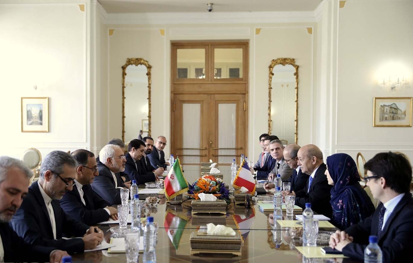 L'image montre une salle de réunion avec une grande table au centre. De chaque côté de la table, plusieurs personnes sont assises, participent à une discussion. Les drapeaux de l'Iran et d'un autre pays sont visibles sur la table. L'environnement est élégant, avec des décorations raffinées et de grandes fenêtres. On peut aussi voir des fleurs au centre de la table. Les participants semblent concentrés et engagés dans la conversation.