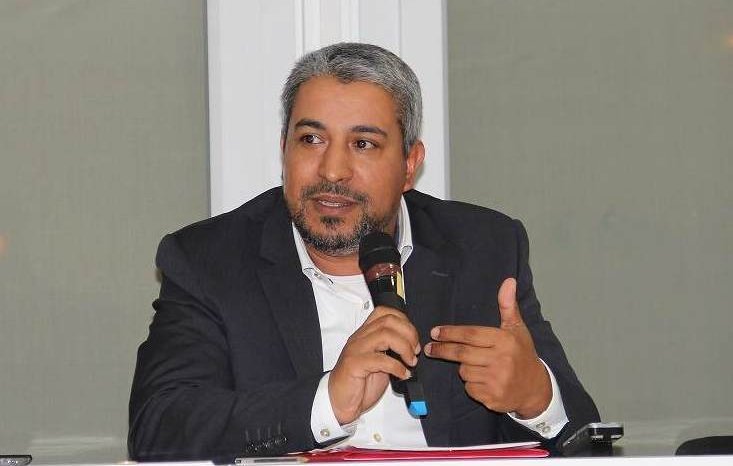 L'image montre un homme assis à une table, tenant un microphone. Il semble engager une discussion ou faire une présentation. Il porte un costume sombre avec une chemise claire et a des cheveux grisonnants. L'environnement autour de lui semble être celui d'une conférence ou d'un événement formel, avec une lumière claire et des fenêtres en arrière-plan.