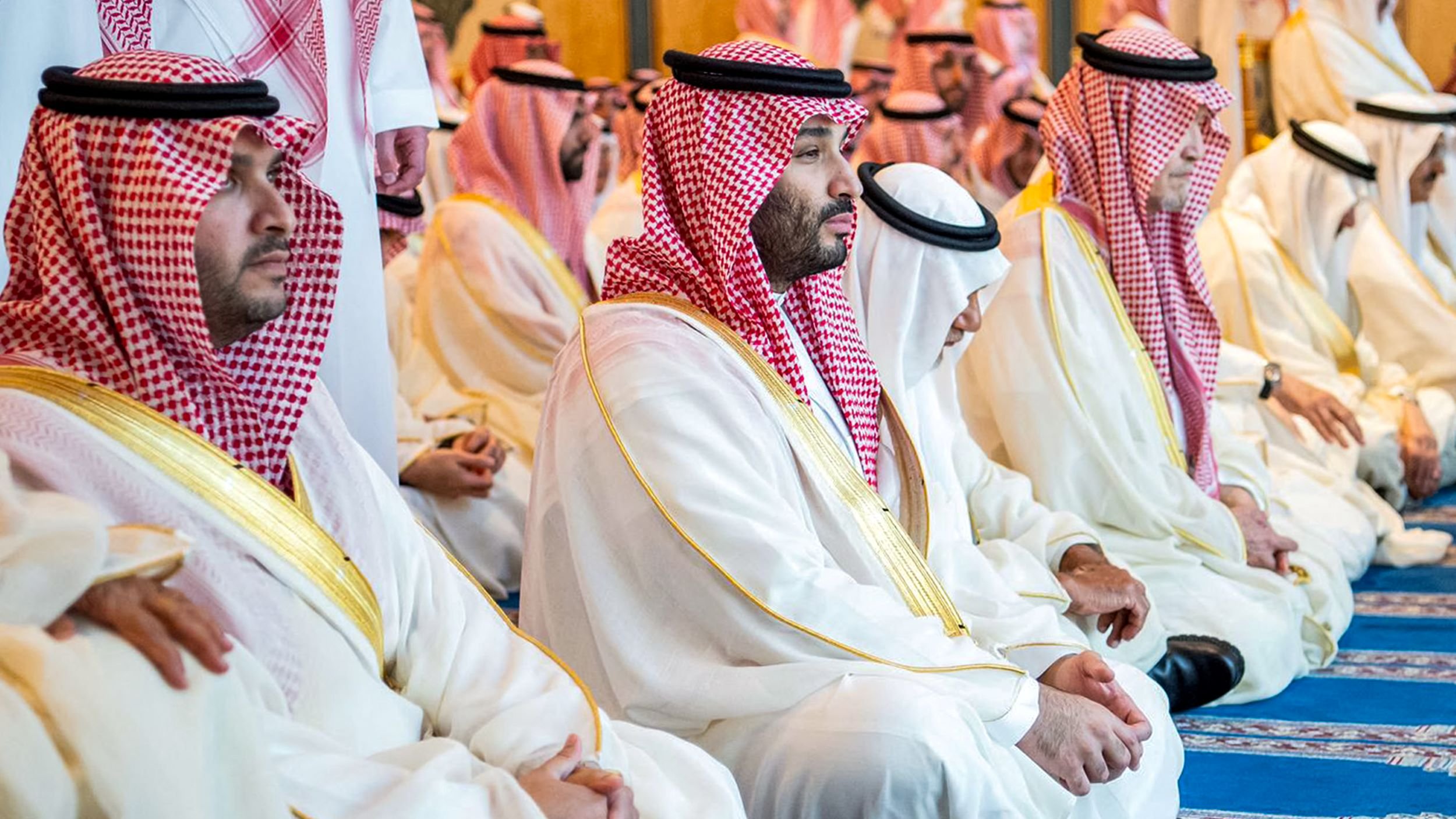 L'image montre un groupe d'hommes assis, probablement en train de prier ou de méditer. Ils portent des vêtements traditionnels saoudiens, comme des thobes blancs et des chapeaux appelés ghutras, souvent ornés de bandes rouges et blanches. L'atmosphère semble empreinte de sérénité et de respect, reflet d'un moment spirituel partagé. Le décor autour d'eux est élégant et peut inclure des éléments architecturaux typiques d'un lieu de culte.
