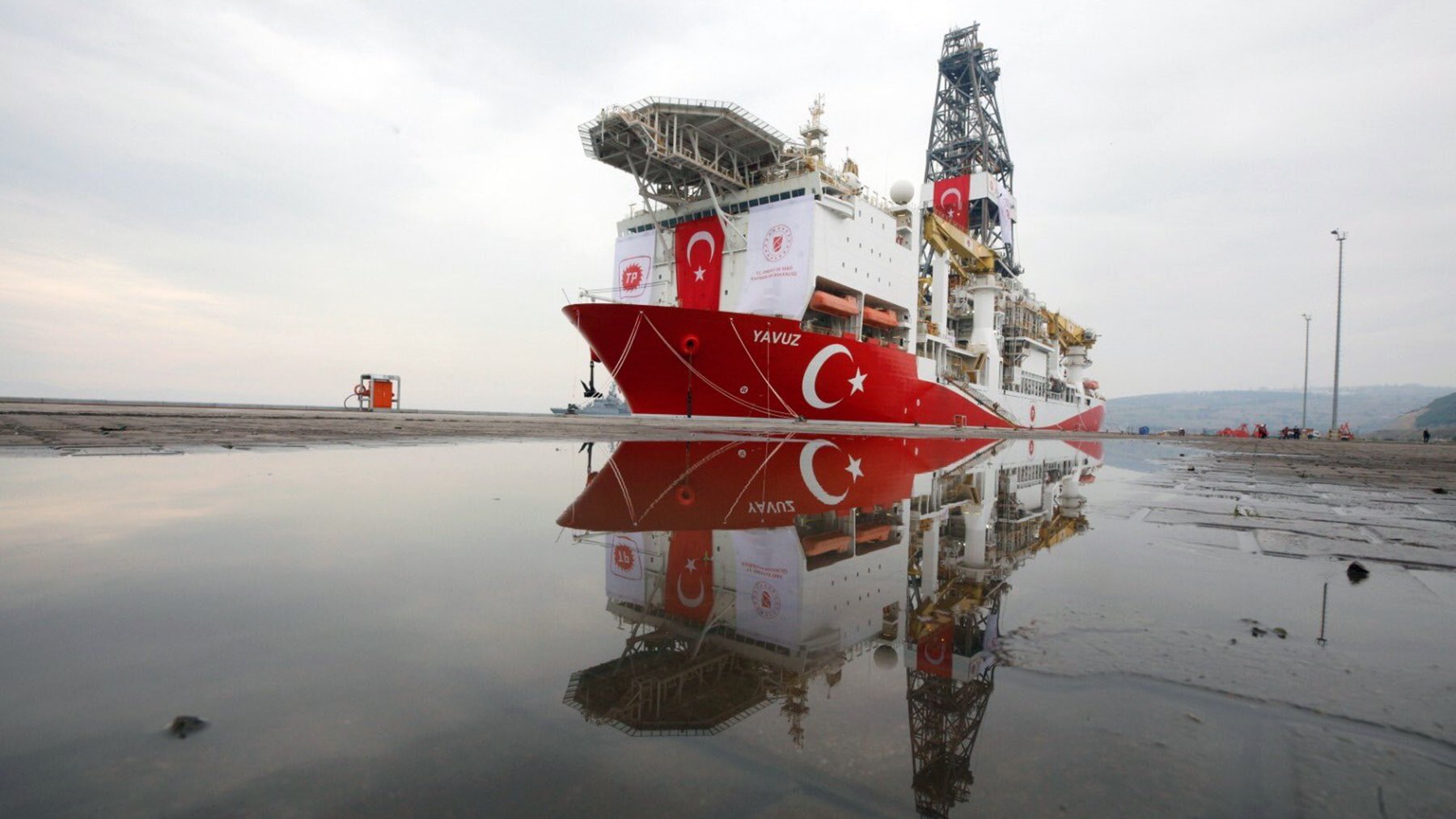 L'image montre un navire de forage maritime, nommé "Yavuz", qui est amarré au port. Le bateau arbore des couleurs dominantes de rouge et blanc, représentant le drapeau turc. On peut également voir des équipements de forage sur le pont du navire. Dans la partie inférieure de l'image, il y a des réflexions du navire dans une flaque d'eau sur le sol. Le ciel en arrière-plan est nuageux, créant une atmosphère calme.