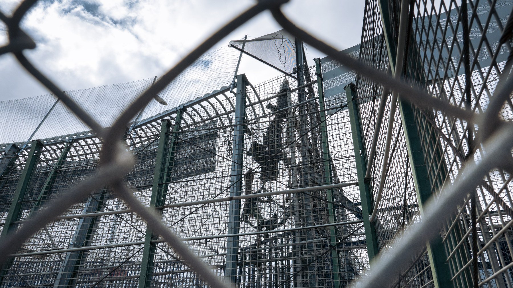 L'image montre une structure en métal, probablement une clôture ou un mur de sécurité, entourée de grillages. On peut apercevoir des barres et une superficie qui semble être une installation sécurisée. Le ciel est nuageux, ce qui donne une atmosphère un peu sombre à la scène. Cette image évoque un environnement restreint, probablement lié à la sécurité ou à la protection.