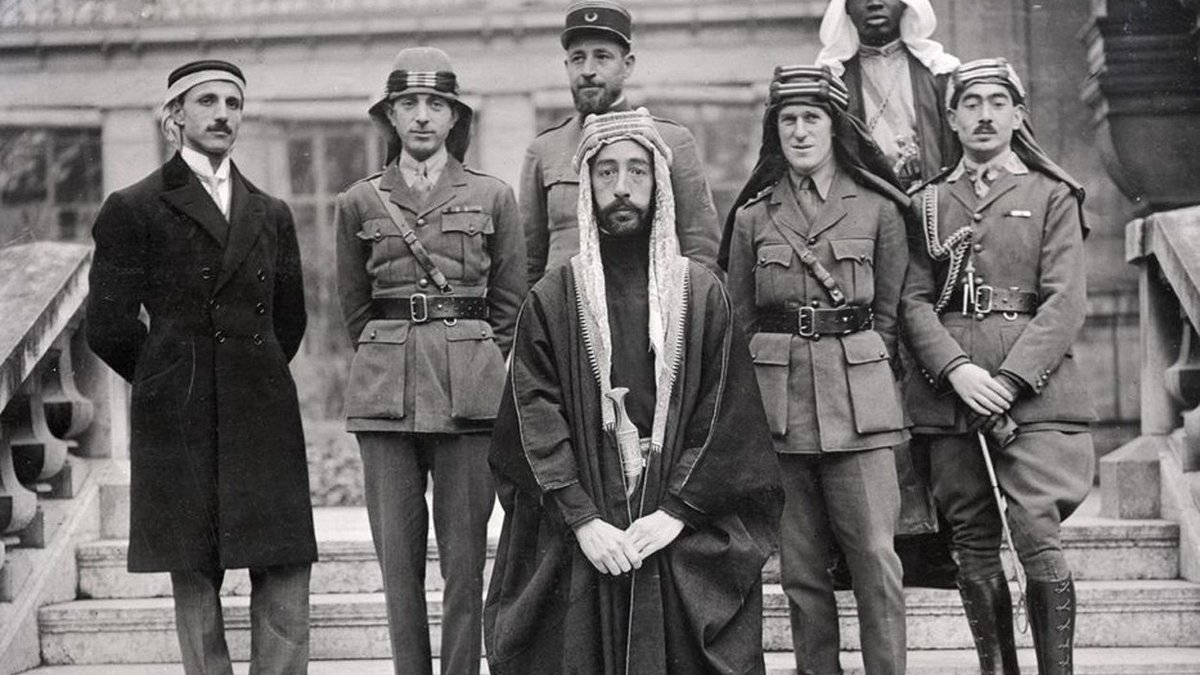 L'image montre un groupe de six hommes posant ensemble. Ils sont vêtus de différentes tenues militaires et traditionnelles. Au centre, un homme en robe traditionnelle, portant une tête couronnée, semble être le principal sujet. À sa gauche, un autre homme en uniforme militaire. Les autres sont également en uniformes variés, indiquant peut-être des origines et des rôles différents. L'arrière-plan montre une architecture qui semble historique. L'ensemble dégage une atmosphère de confluence culturelle et de collaboration.