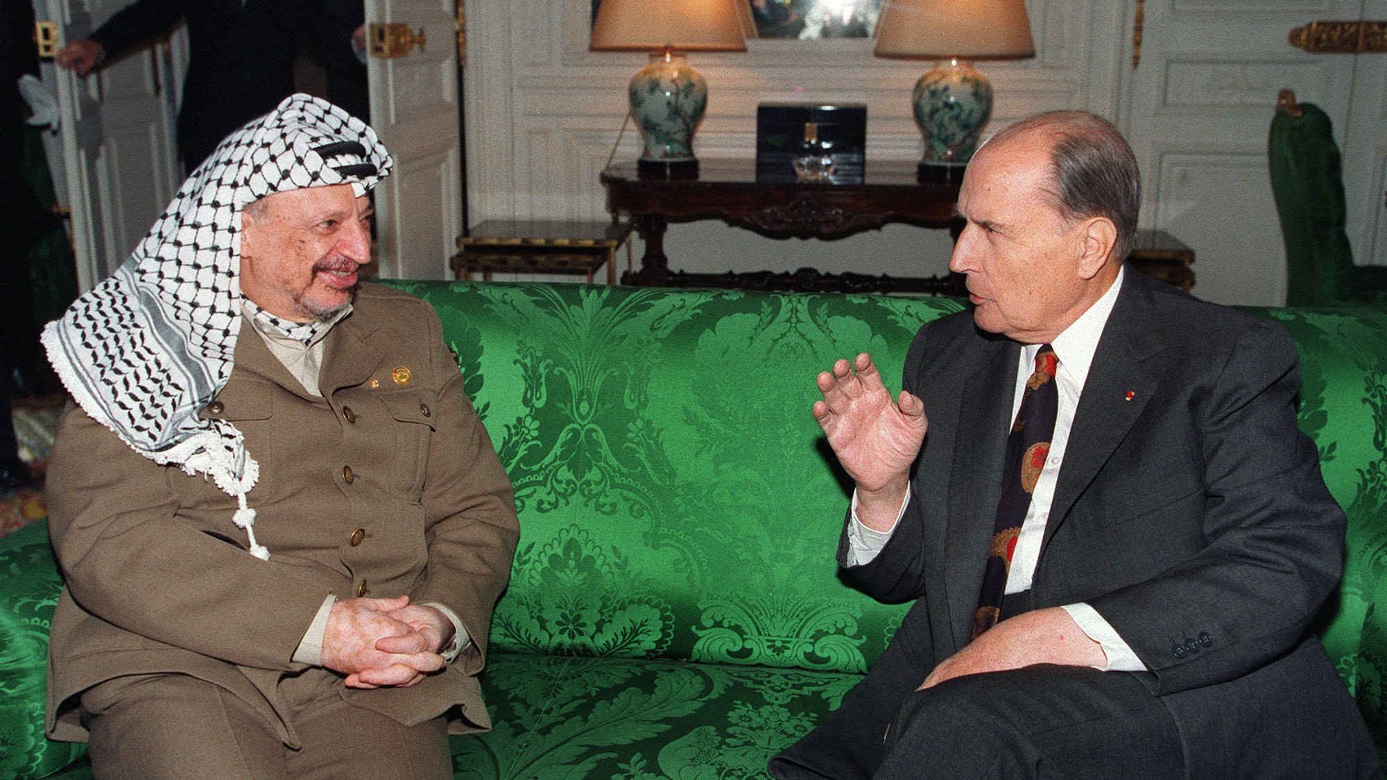 L'image montre deux hommes assis sur un canapé vert décoré. L'un des hommes porte un costume beige avec un keffieh, tandis que l'autre est en costume sombre avec une cravate à motifs. Ils semblent engagés dans une conversation animée. En arrière-plan, on aperçoit des éléments de mobilier et une lumière douce, suggérant un cadre intérieur élégant.