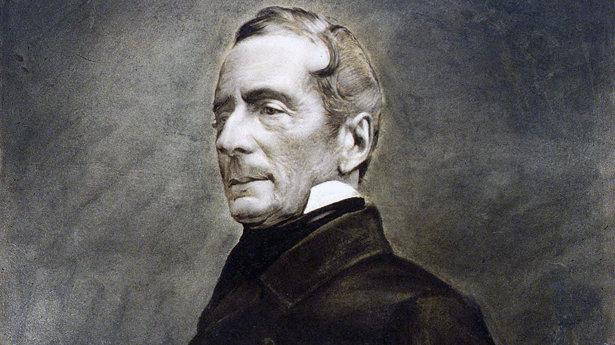 L'image montre un portrait en noir et blanc d'un homme d'âge moyen, vêtu d'un manteau sombre. Son expression est sérieuse et réfléchie, avec des traits du visage bien définis. Il a les cheveux légèrement ondulés, peignés vers l'arrière, et il porte un col montant. L'arrière-plan est flou, mettant en valeur le sujet du portrait. L'ensemble a un aspect classique et intemporel, caractéristique des portraits du 19ème siècle.