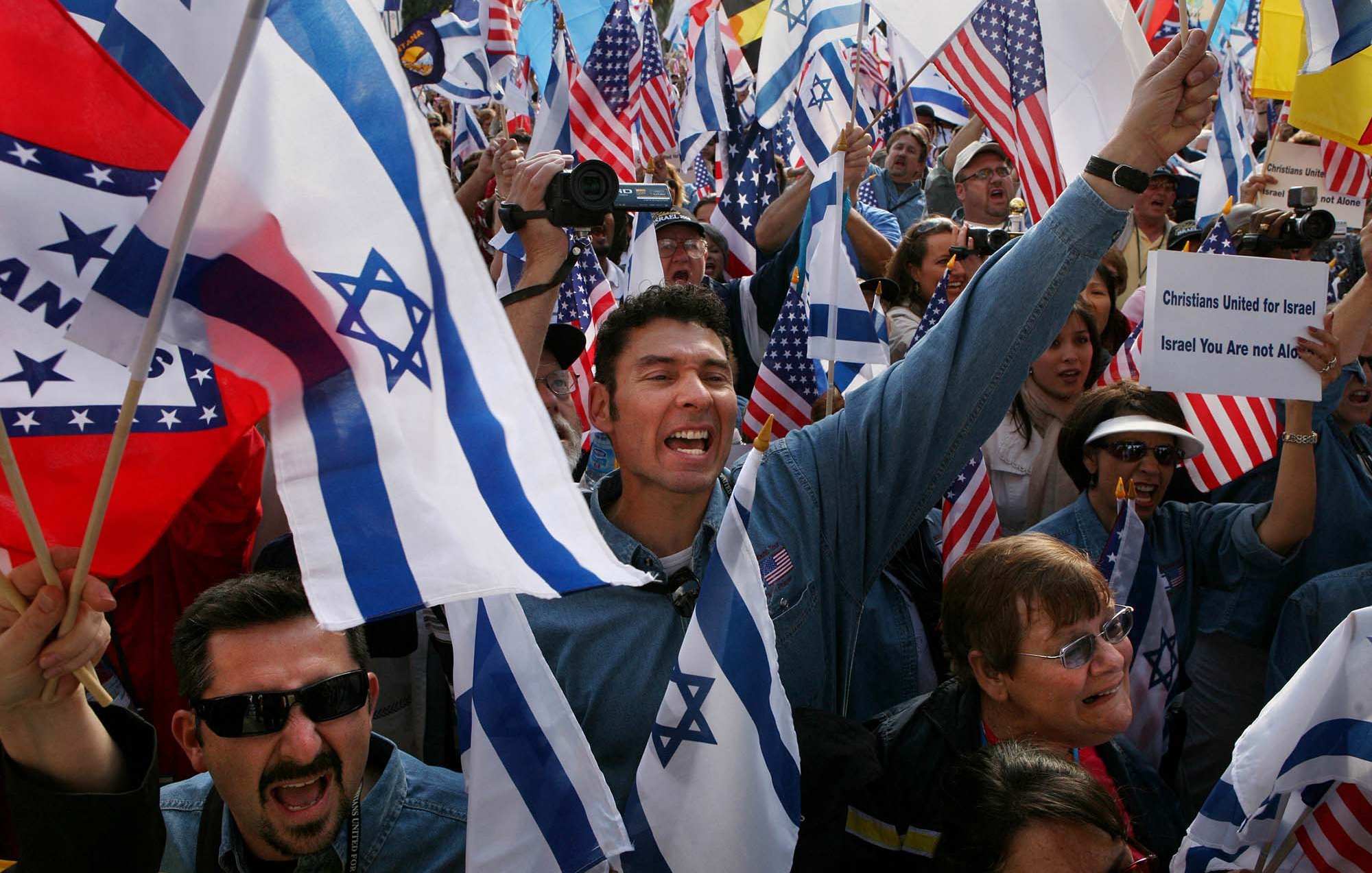 L'image montre une manifestation avec des participants brandissant des drapeaux israéliens et américains. Les gens semblent très engagés et animés, certains lèvent les bras en l'air et expriment leur enthousiasme, tandis que d'autres portent des expressions de concentration ou de soutien. Il y a un mélange de différentes personnes dans la foule, suggérant une diversité au sein de ce rassemblement.